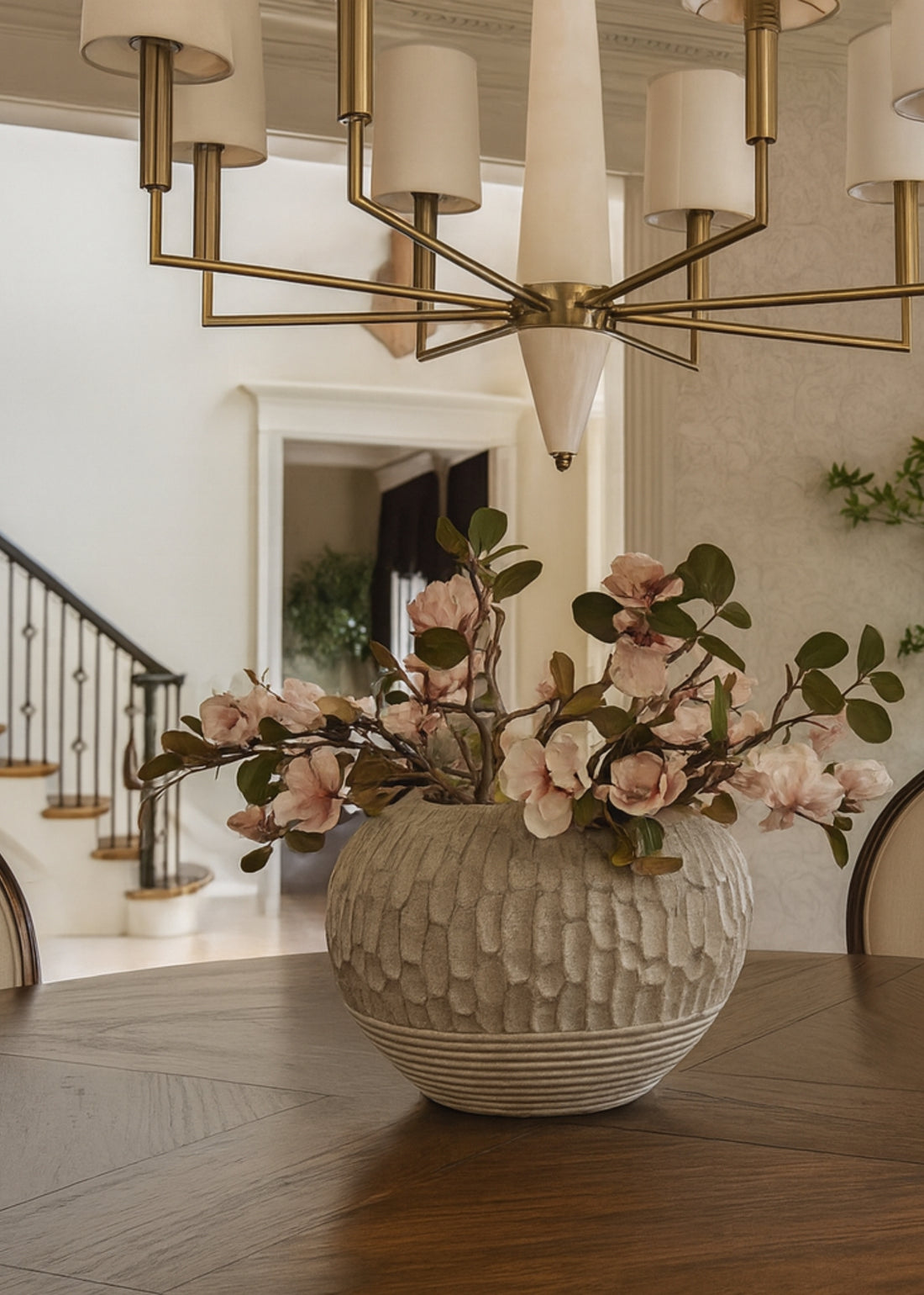 Decorative vase with pink magnolia flowers on a table in a room with a chandelier.