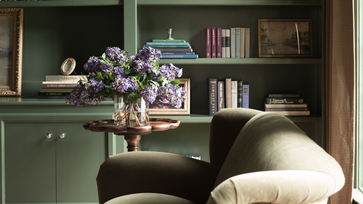 Decorative interior with a green armchair, bookshelf, and vase of purple flowers.