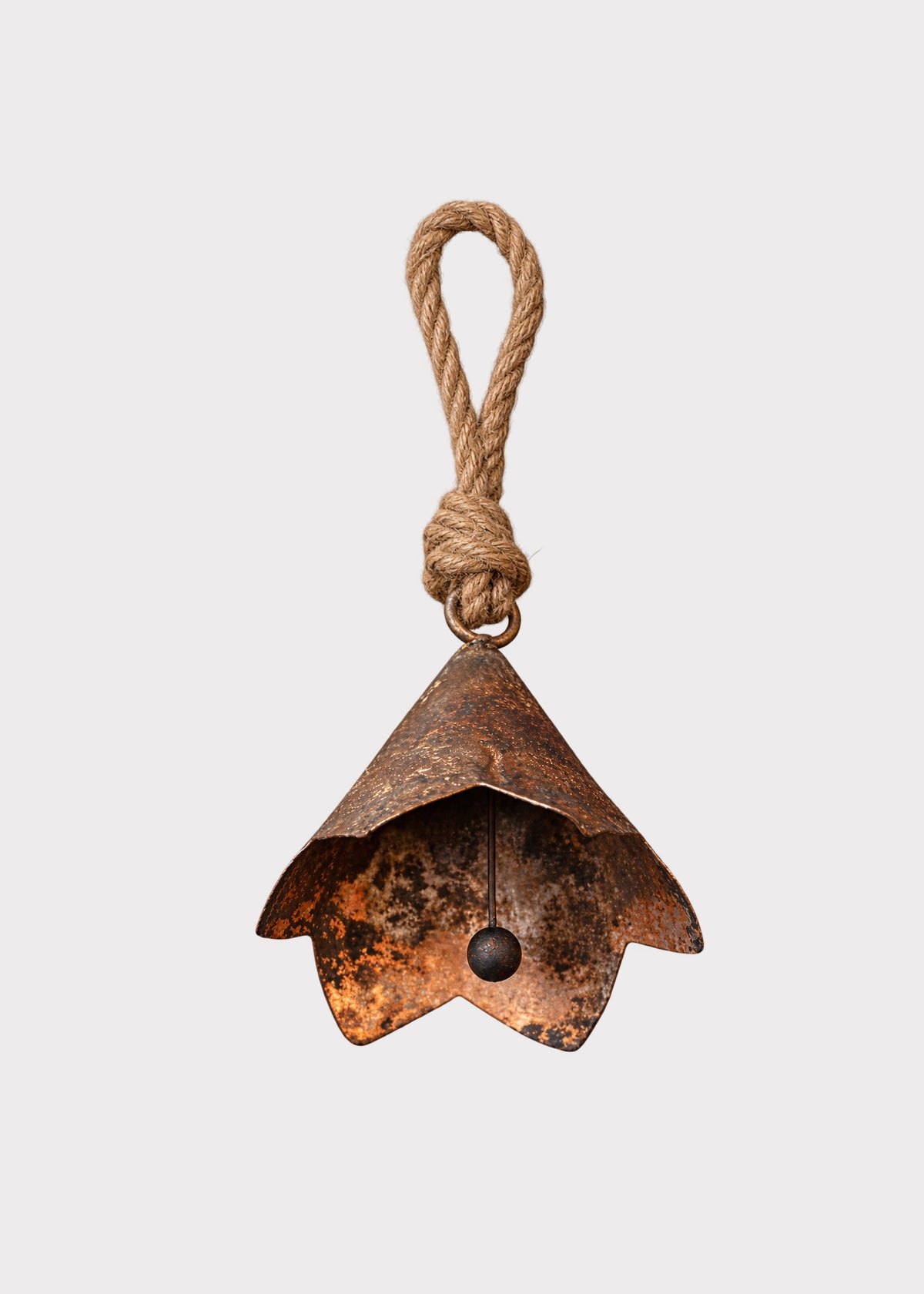 A rustic, handcrafted metal bell ornament with a weathered copper-brown patina and a natural jute rope loop at the top, shown against a solid light gray background.