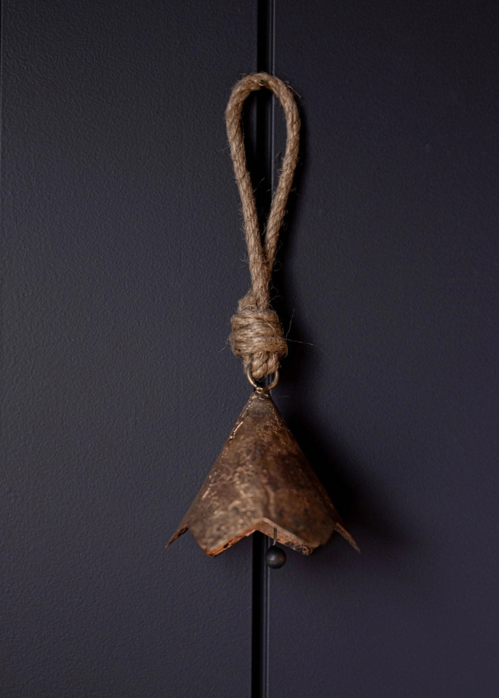 A rustic brown metal bell ornament hanging from a thick natural jute rope loop, set against a dark charcoal gray background.