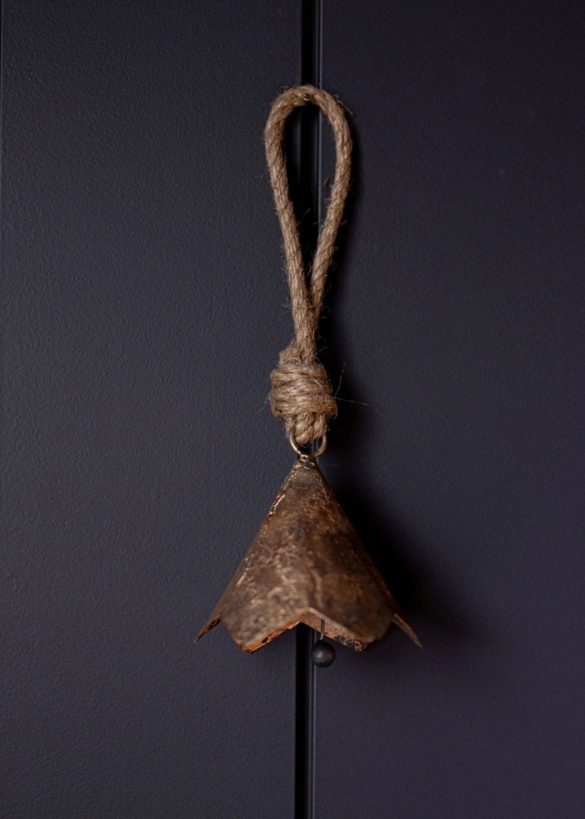 A rustic brown metal bell ornament hanging from a thick natural jute rope loop, set against a dark charcoal gray background.