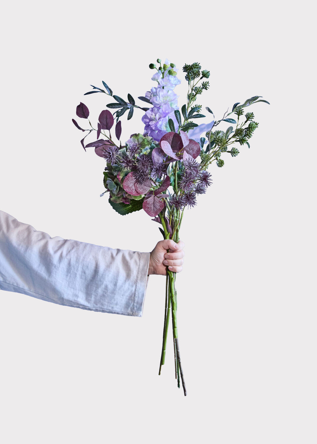 A hand in a white long-sleeve shirt holds a fake flower bouquet with purple and green botanicals 