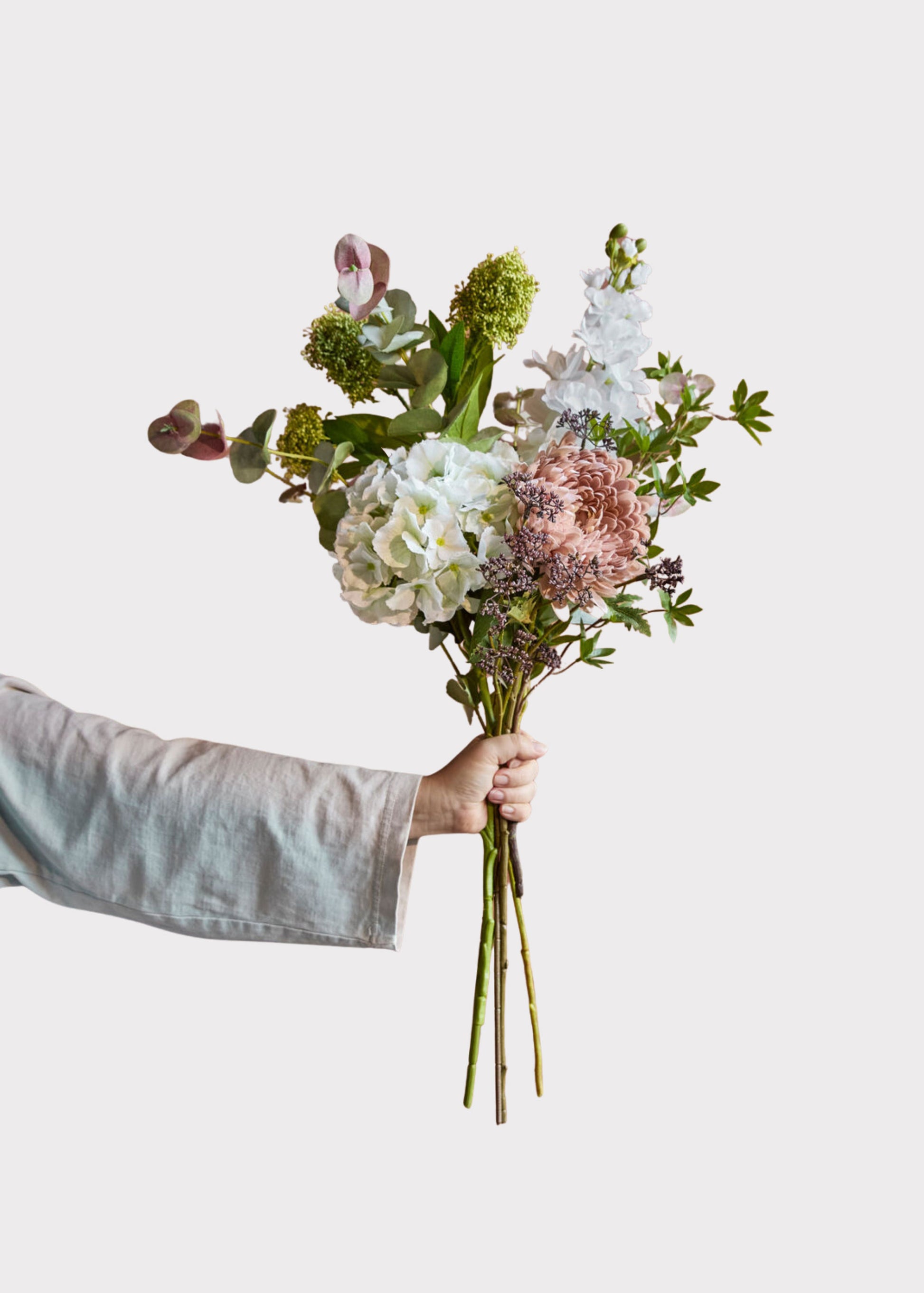 A hand holding a faux flower bouquet with white hydrangeas, dusty pink blooms, and green foliage