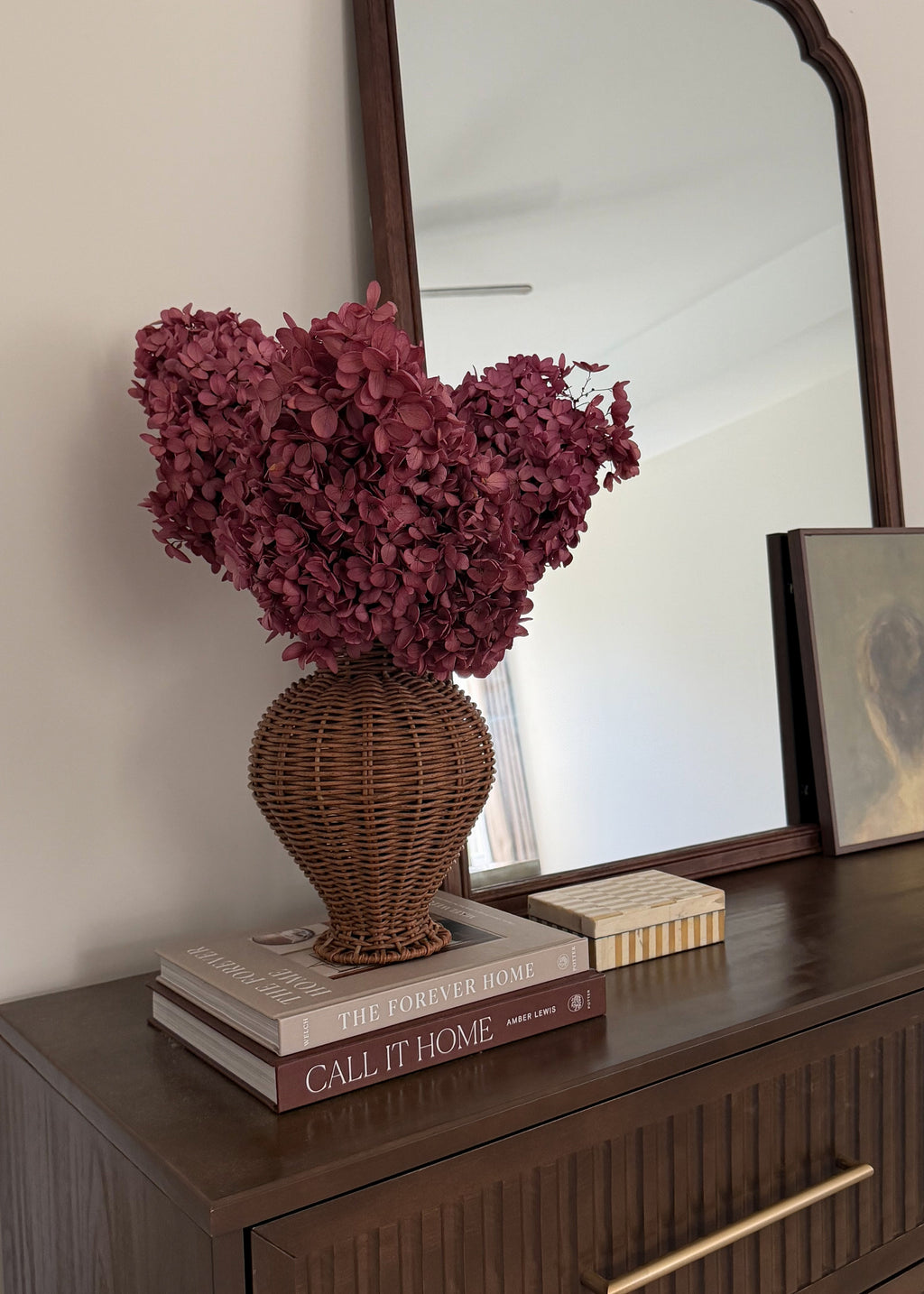 A decorative arrangement featuring a large bouquet of burgundy dried hydrangeas in a brown woven wicker vase, displayed on a dark wood dresser next to books and a mirror.