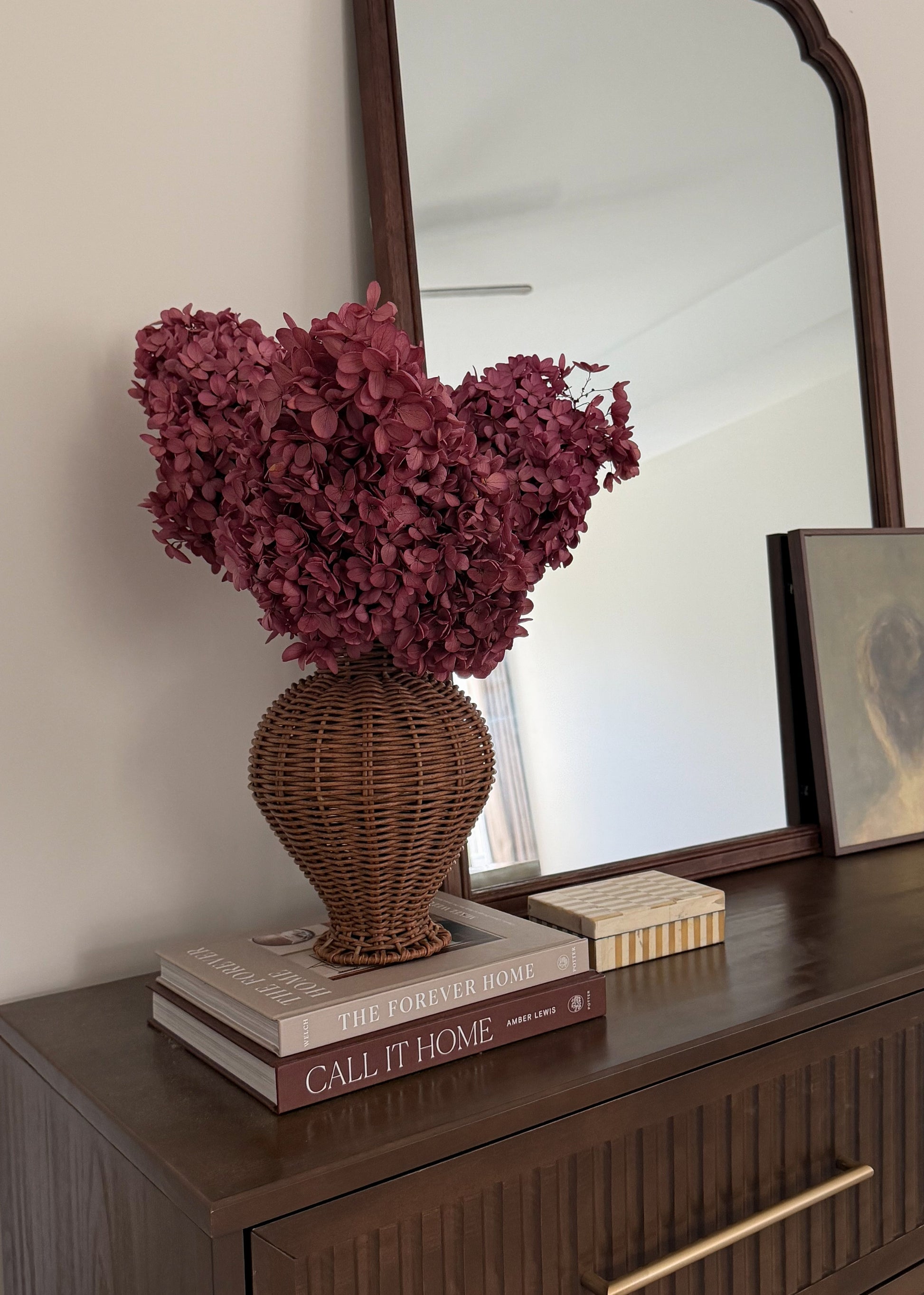 A decorative arrangement featuring a large bouquet of burgundy dried hydrangeas in a brown woven wicker vase, displayed on a dark wood dresser next to books and a mirror.