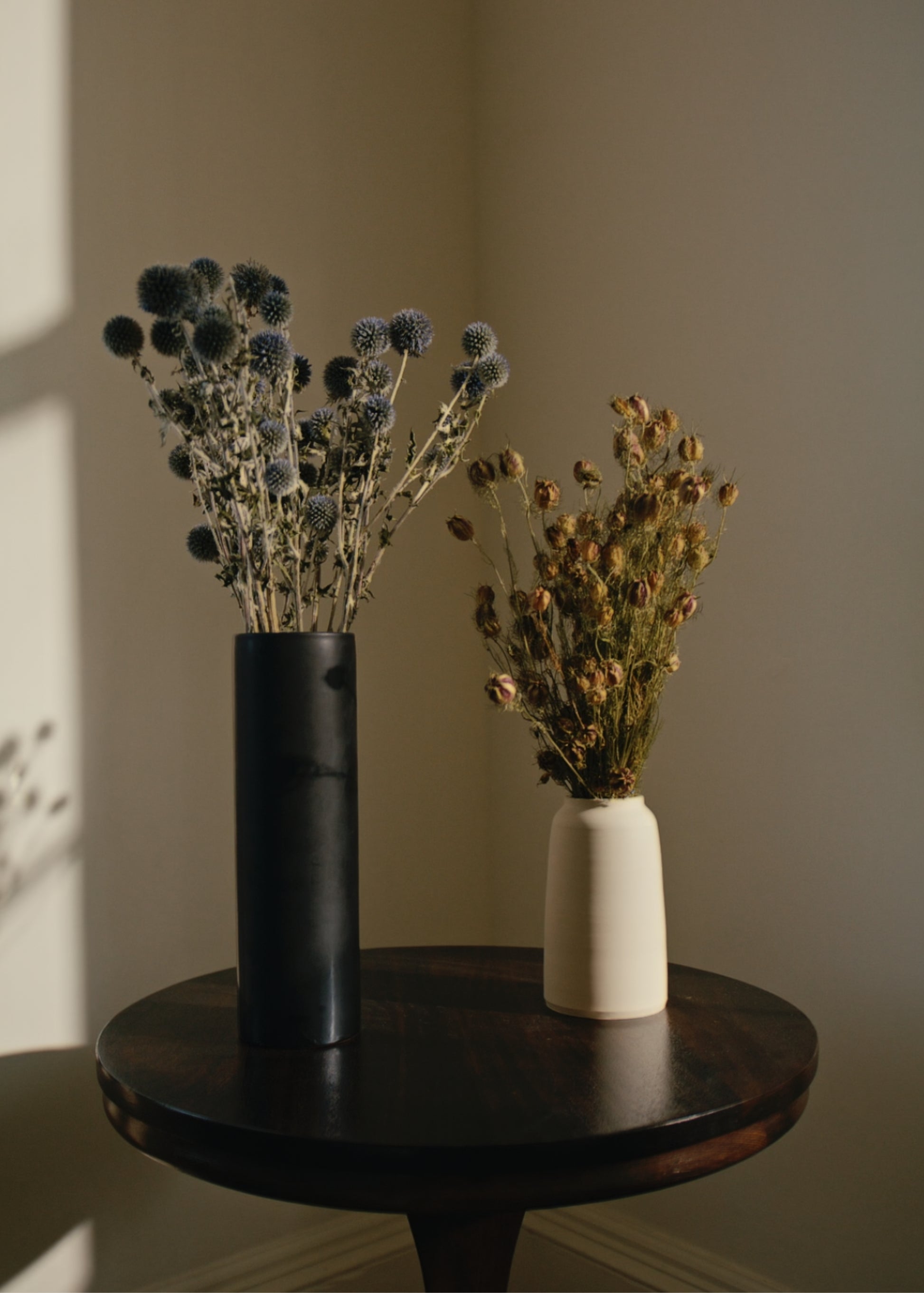 Two vases with dried flowers on a wooden table against a plain wall.