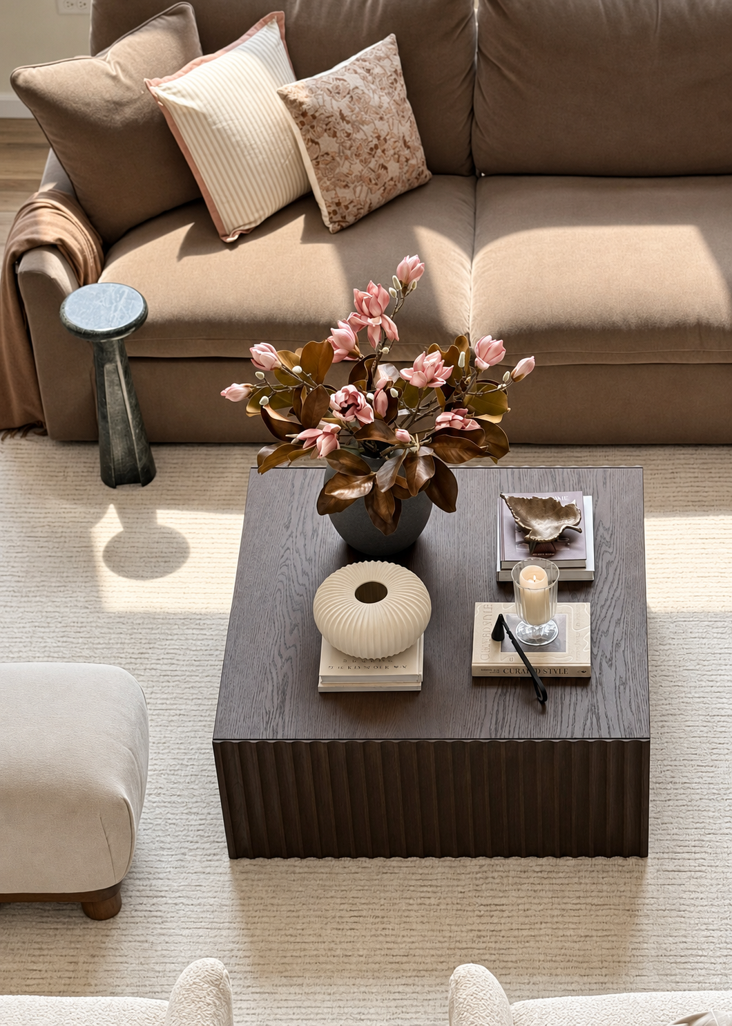 A gray ceramic vase filled with dried pink and brown magnolias on a dark wooden table in a cozy living room setting with a stone fireplace and white cabinets in the background.