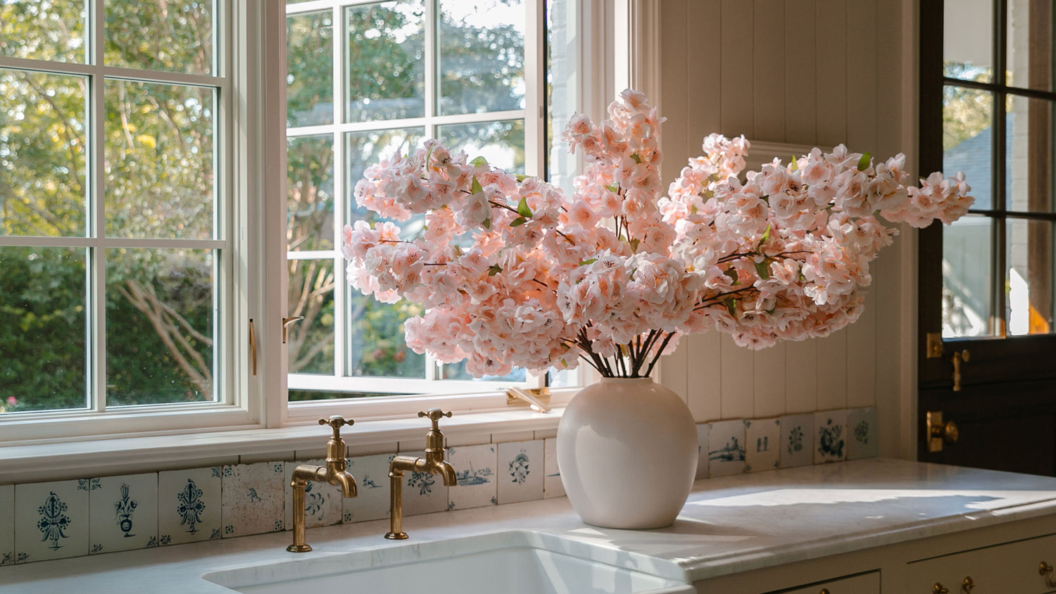 pink cherry blossoms arranged in stoneware vase by open window