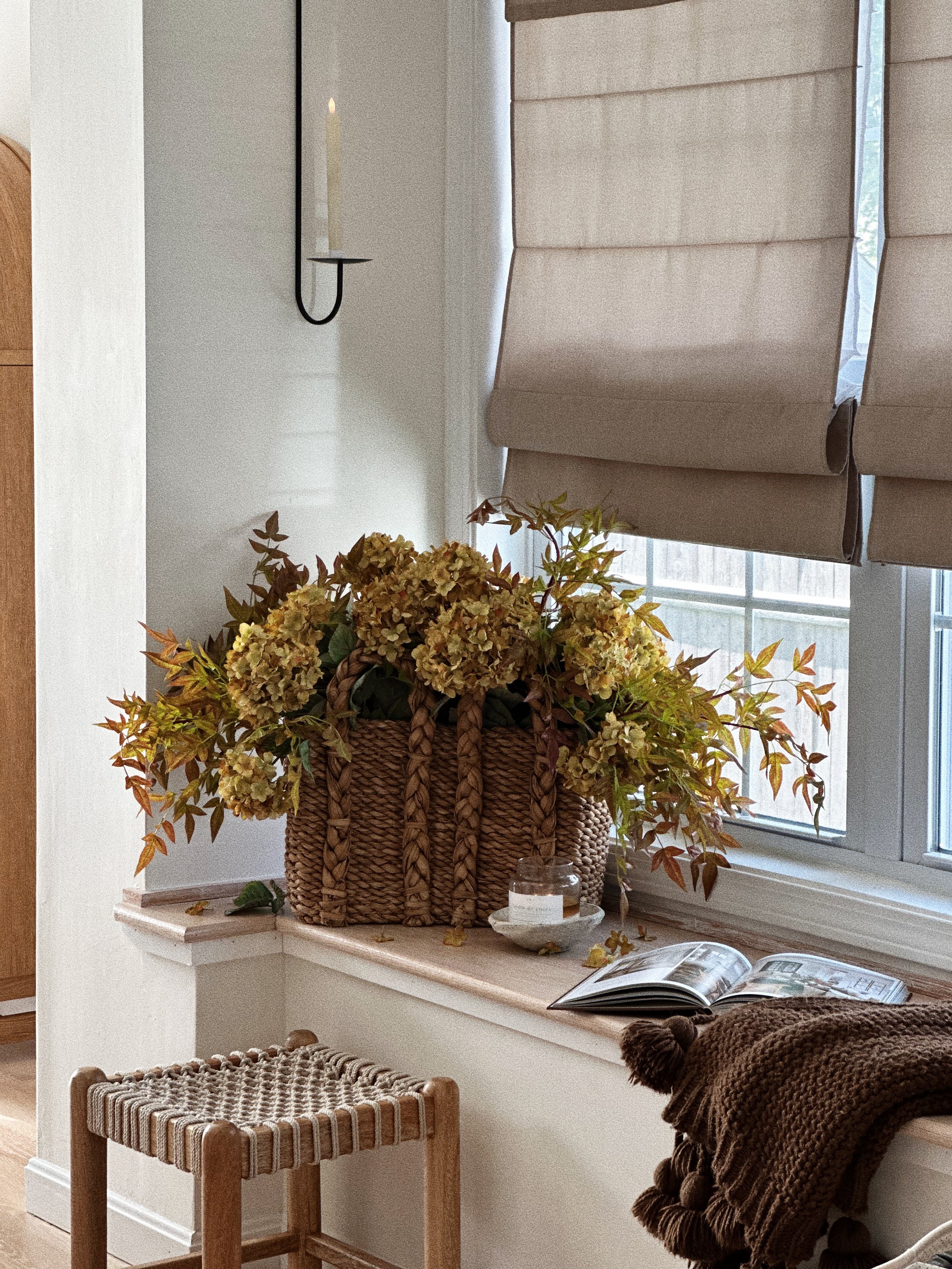 A styled interior corner with a large woven basket filled with dried yellow-green flowers on a windowsill, next to a window with beige roller shades, a small woven stool, and a brown knit throw blanket.