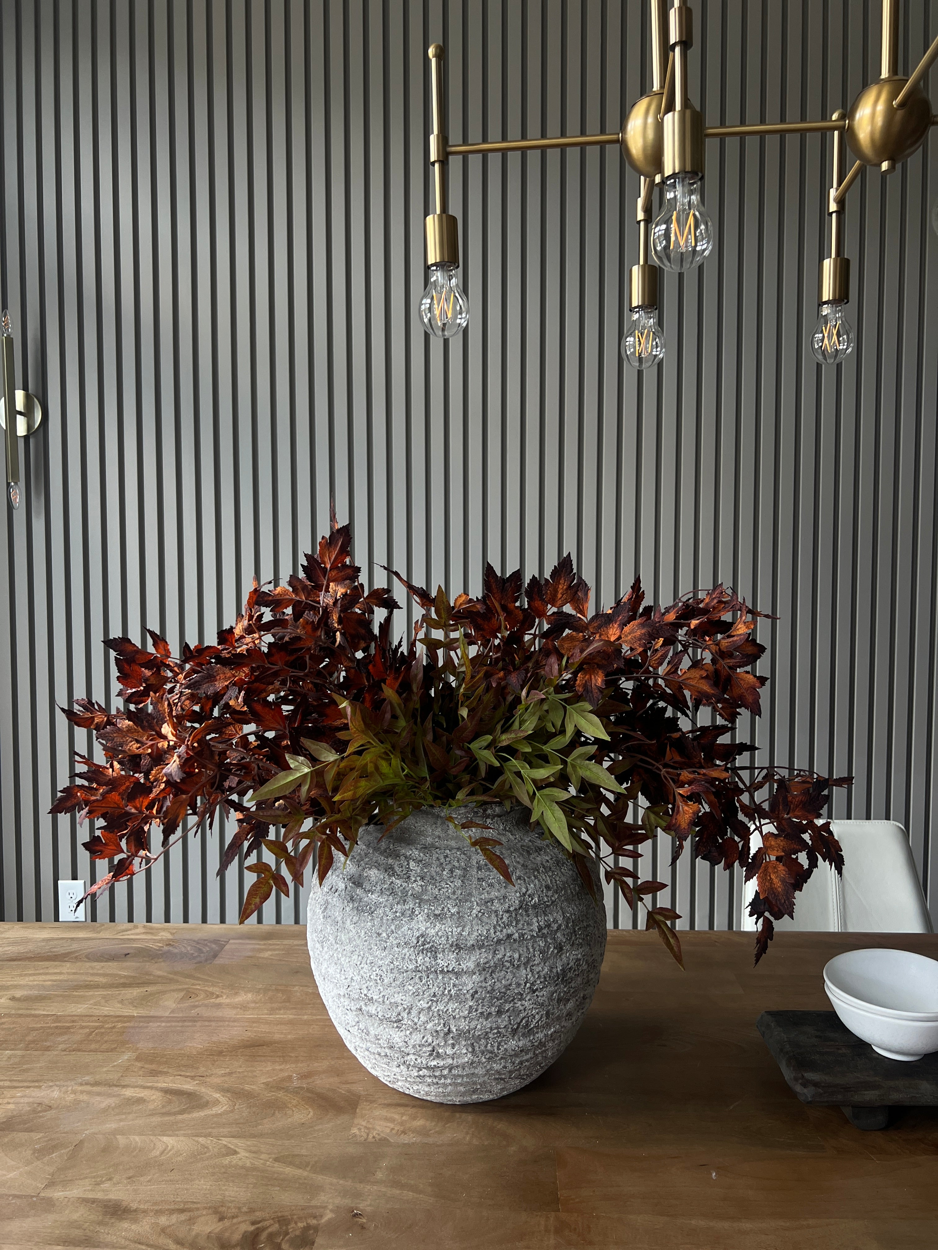 A decorative arrangement of dark burgundy and green artificial foliage in a round textured gray pot, placed on a wooden floor against a gray slatted wall.