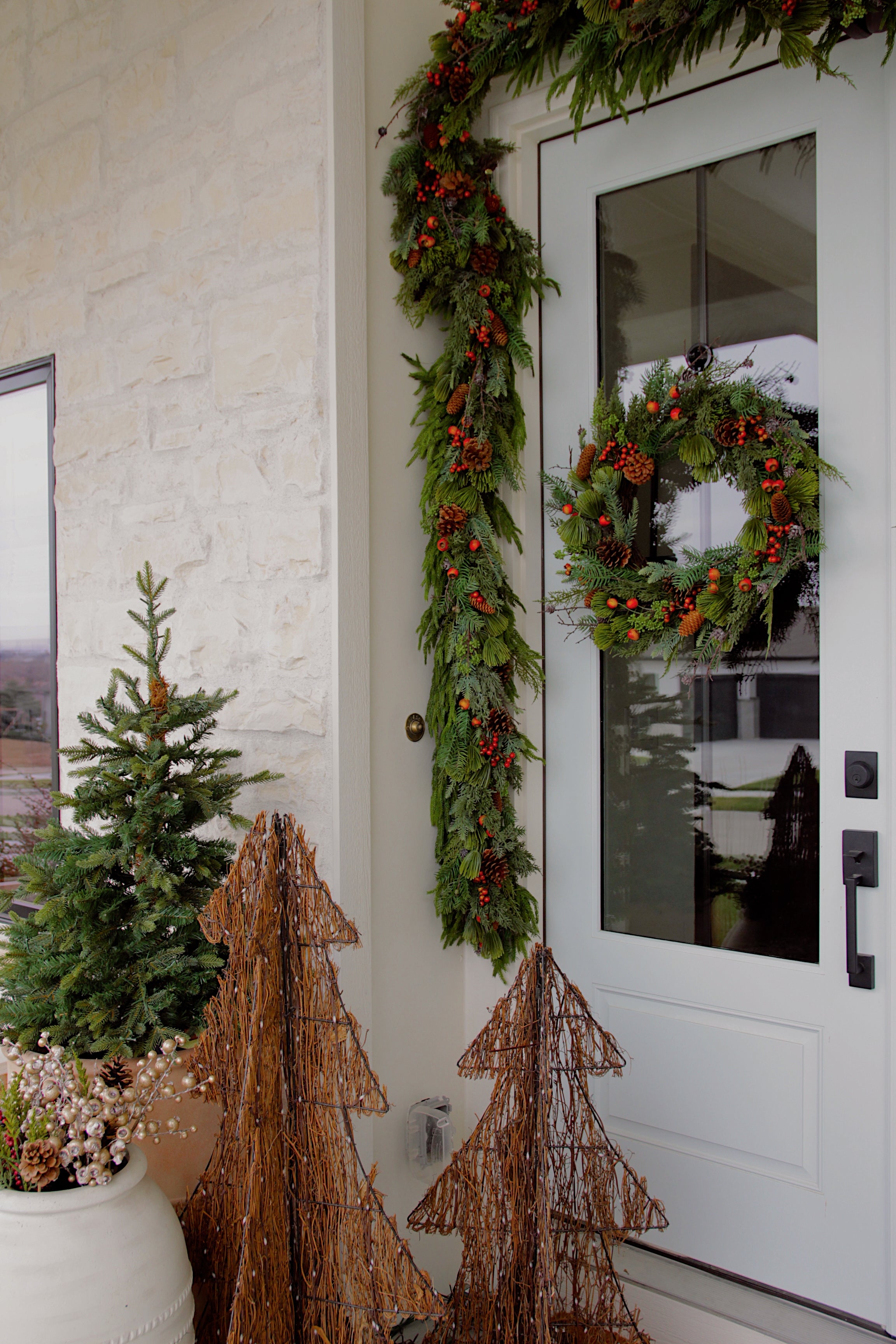 A white front door decorated with a green garland and berry wreath, with matching holiday greenery and wire tree decorations on a porch beside a white brick wall.