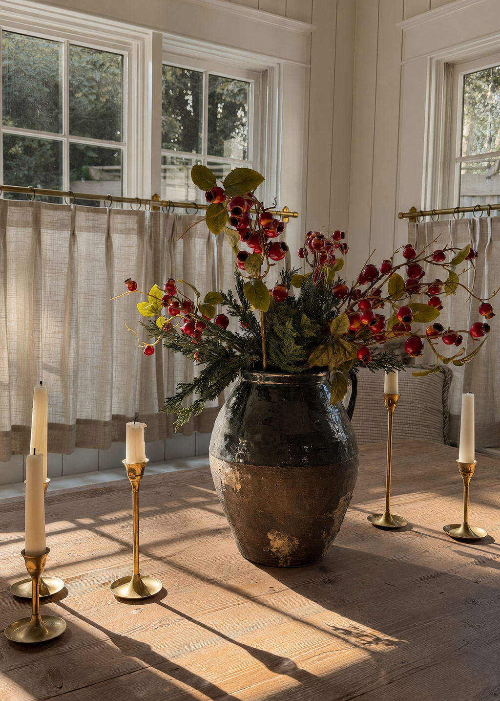 A rustic dark brown ceramic vase filled with a winter floral arrangement of red crabapples and cedar, set on a wooden table in a sunlit room with candles.