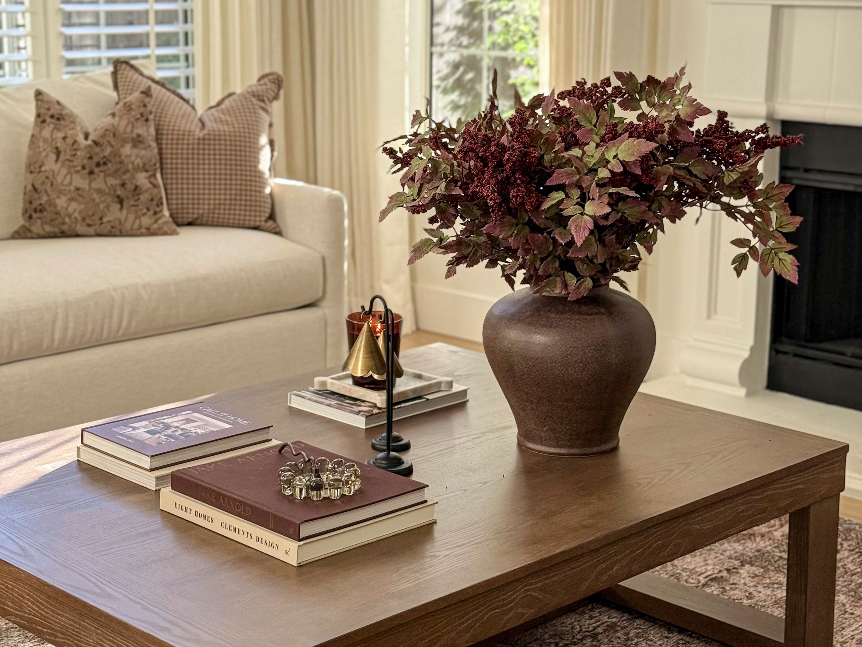 A rustic brown ceramic vase filled with burgundy and green artificial foliage, placed on a wooden coffee table in a styled living room next to a cream sofa.