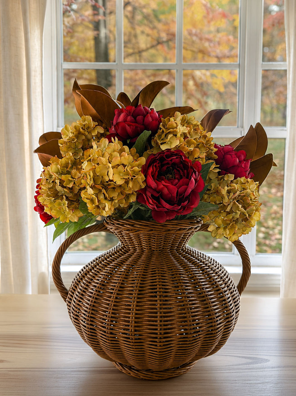 A decorative wicker basket vase filled with artificial autumn flowers, including yellow hydrangeas, red peonies, and brown leaves, set on a light wood table in front of a window with a fall view outside.
