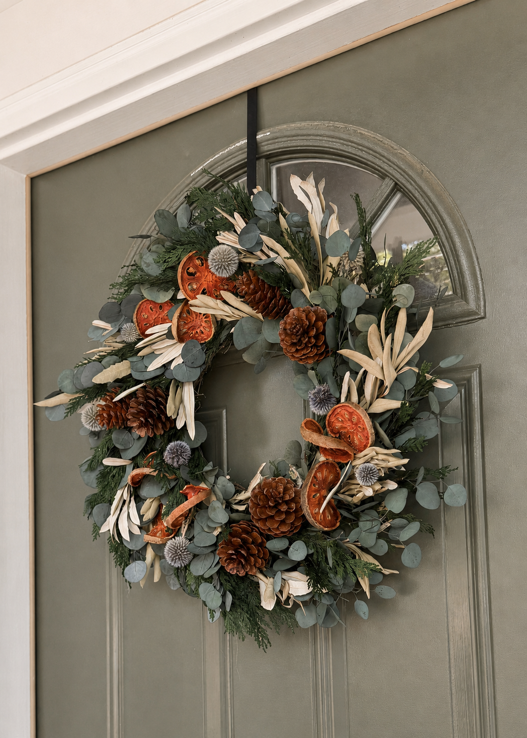 A decorative autumn wreath hanging on a muted sage green front door. The wreath features green eucalyptus foliage, pine cones, dried orange slices, and cream-colored leaves.