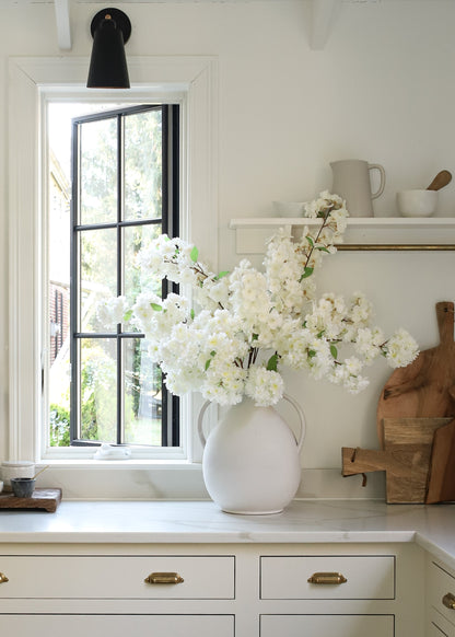 White vase with white cherry blossoms on a kitchen counter next to a window