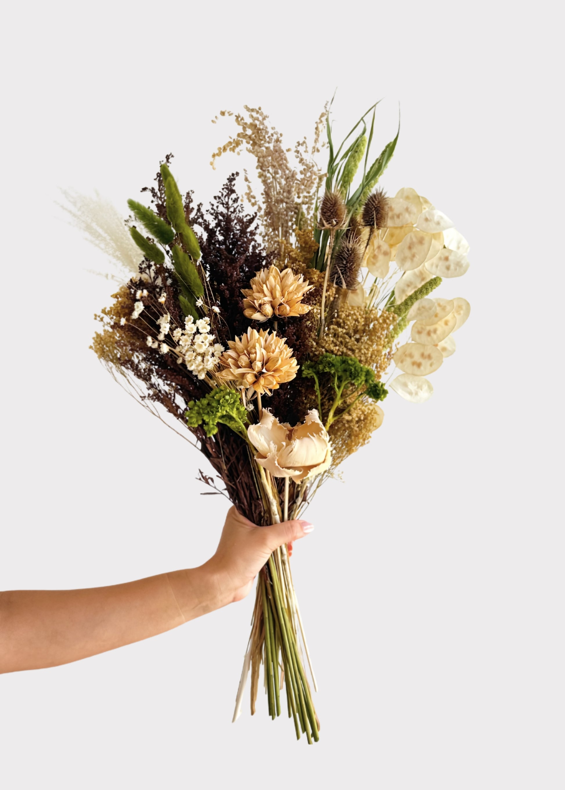 A hand holding a dried flower bouquet with beige, cream, brown, and green botanical elements against a plain light gray background.