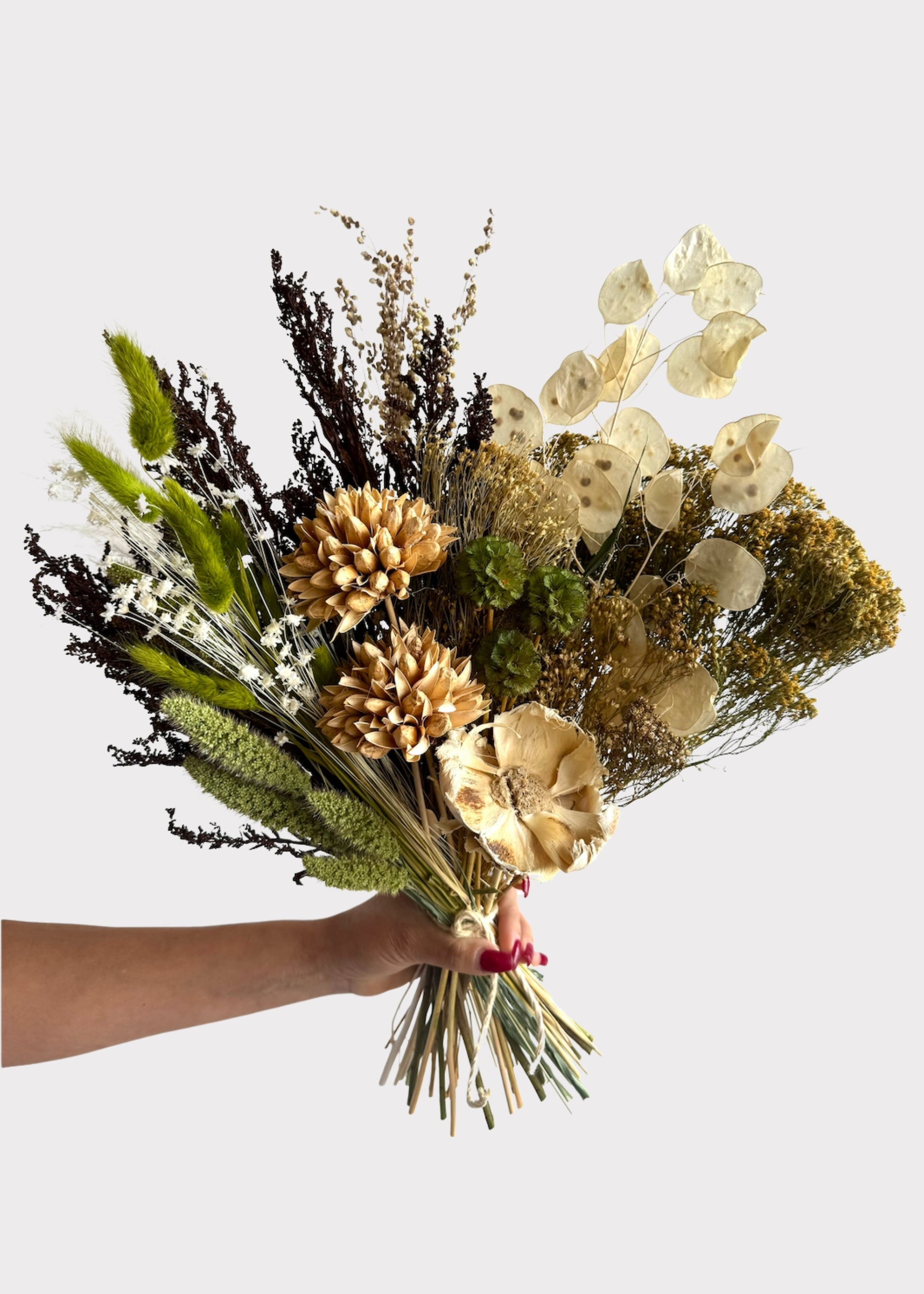 A hand holding a dried flower bouquet with beige, cream, brown, and green botanical elements against a plain light gray background.