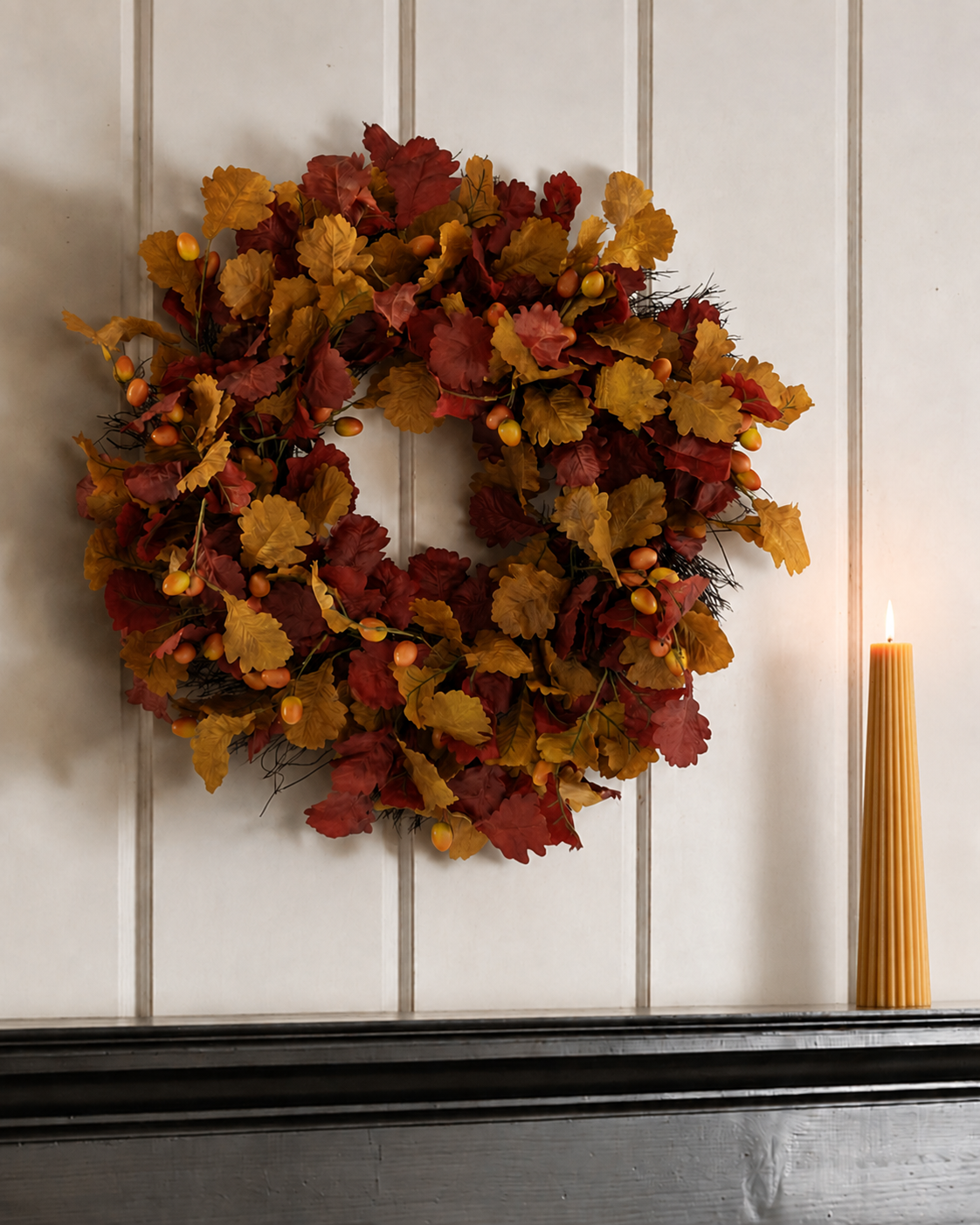 An autumn leaf wreath with red, orange, and yellow faux leaves and small berries, hanging on a white paneled wall above a dark mantel, with a lit candle on the right.