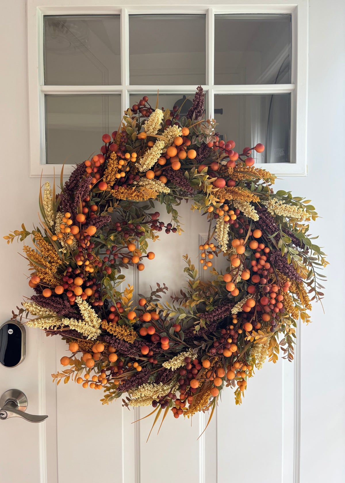An autumn-themed artificial wreath with orange berries, yellow wheat-like accents, and dark purple foliage, hanging on a white paneled front door.