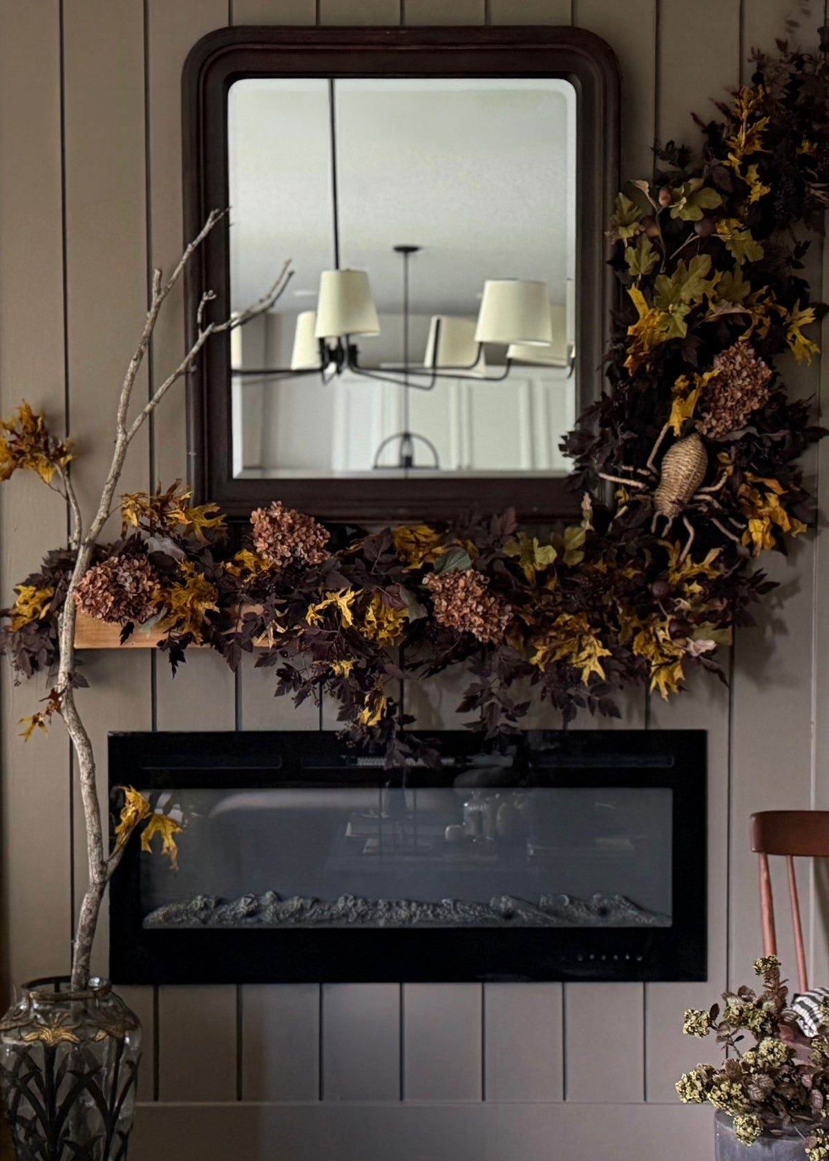 An indoor fireplace mantel styled with a large dark wood mirror, a dark fireplace insert, and a decorative cimicifuga garland with preserved autumn leaves and faux dried hydrangeas in brown and gold tones.