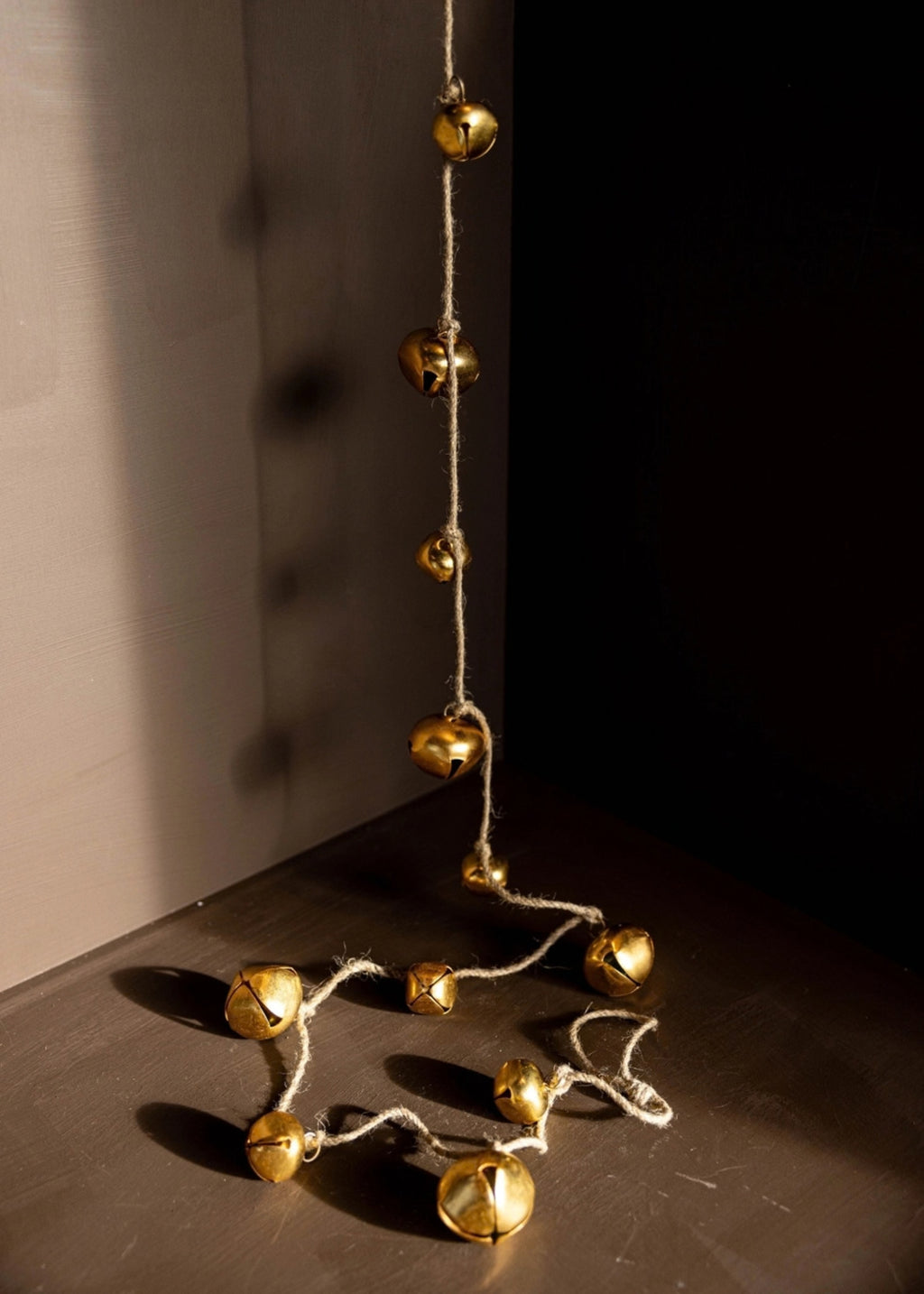 A string of small metallic gold bells hanging from a rope, draped across a dark surface in a dimly lit corner with strong sunlight and shadows.