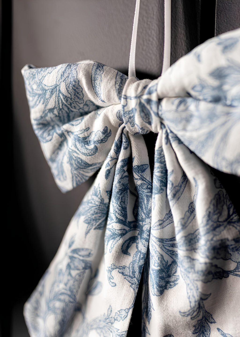 A close-up of a fabric bow accessory with a blue floral toile pattern on an off-white background, tied in a knot and hanging against a dark gray background.