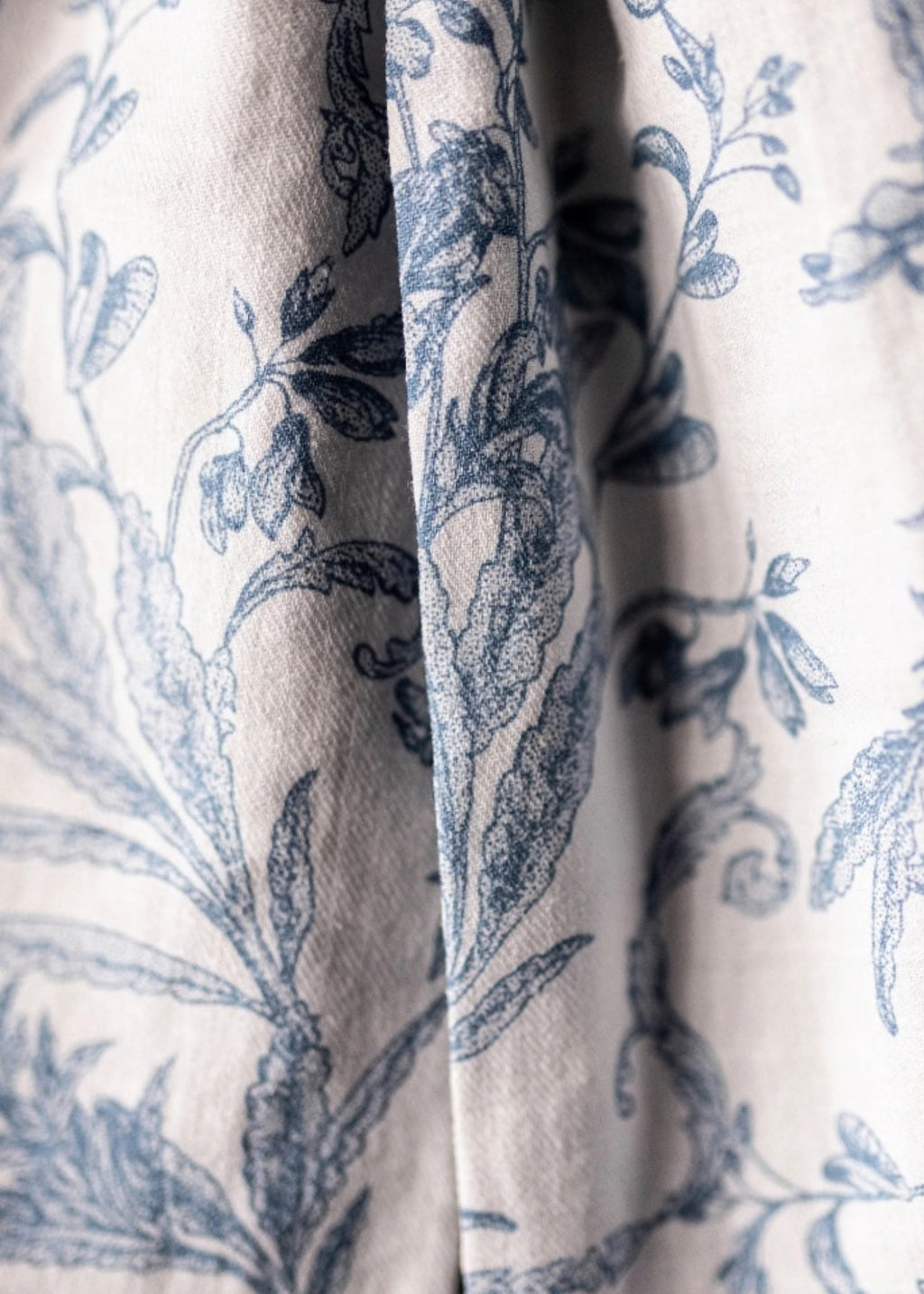 Close-up of a draped fabric with a blue botanical toile pattern on a warm off-white background.