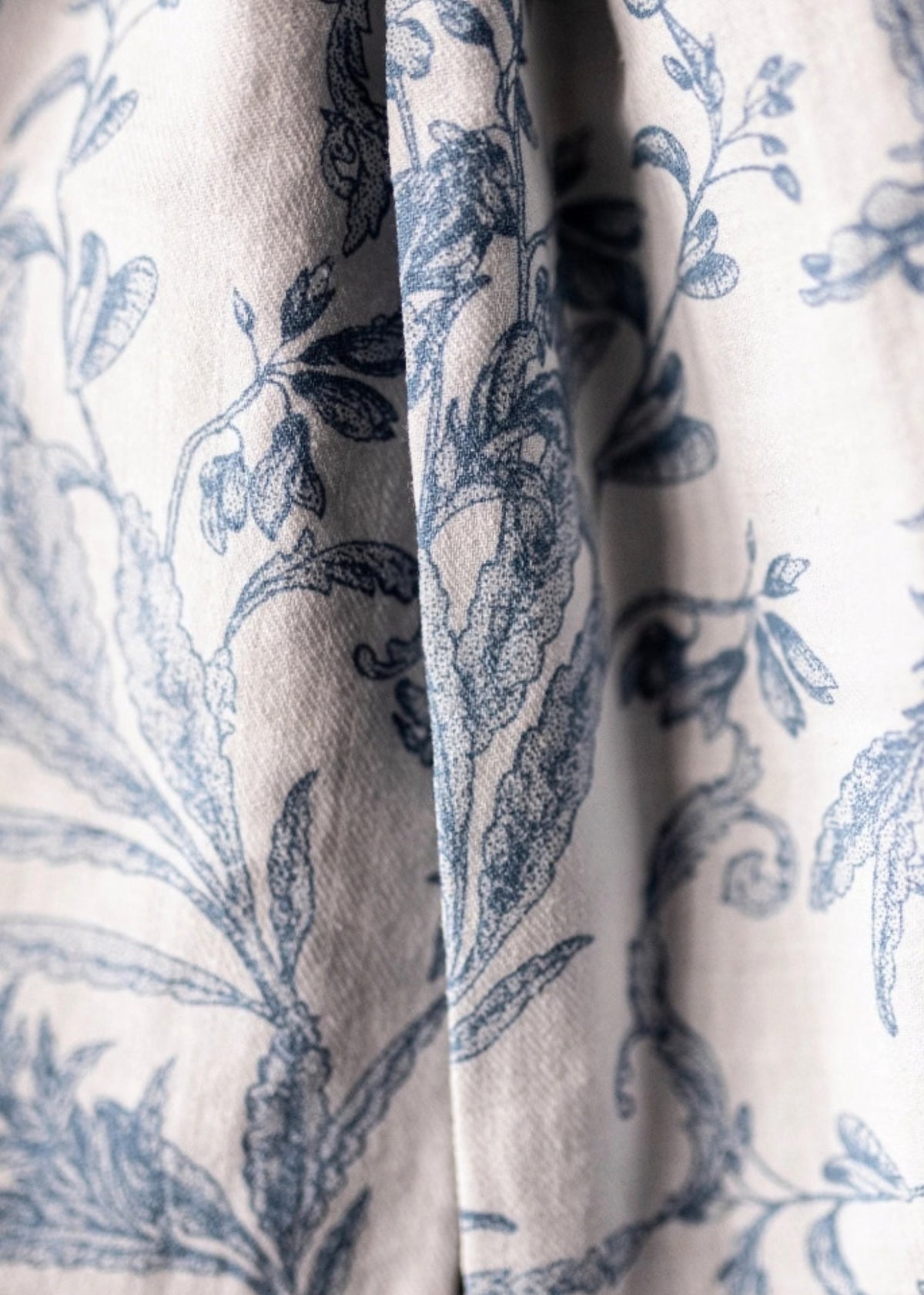 Close-up of a draped fabric with a blue botanical toile pattern on a warm off-white background.