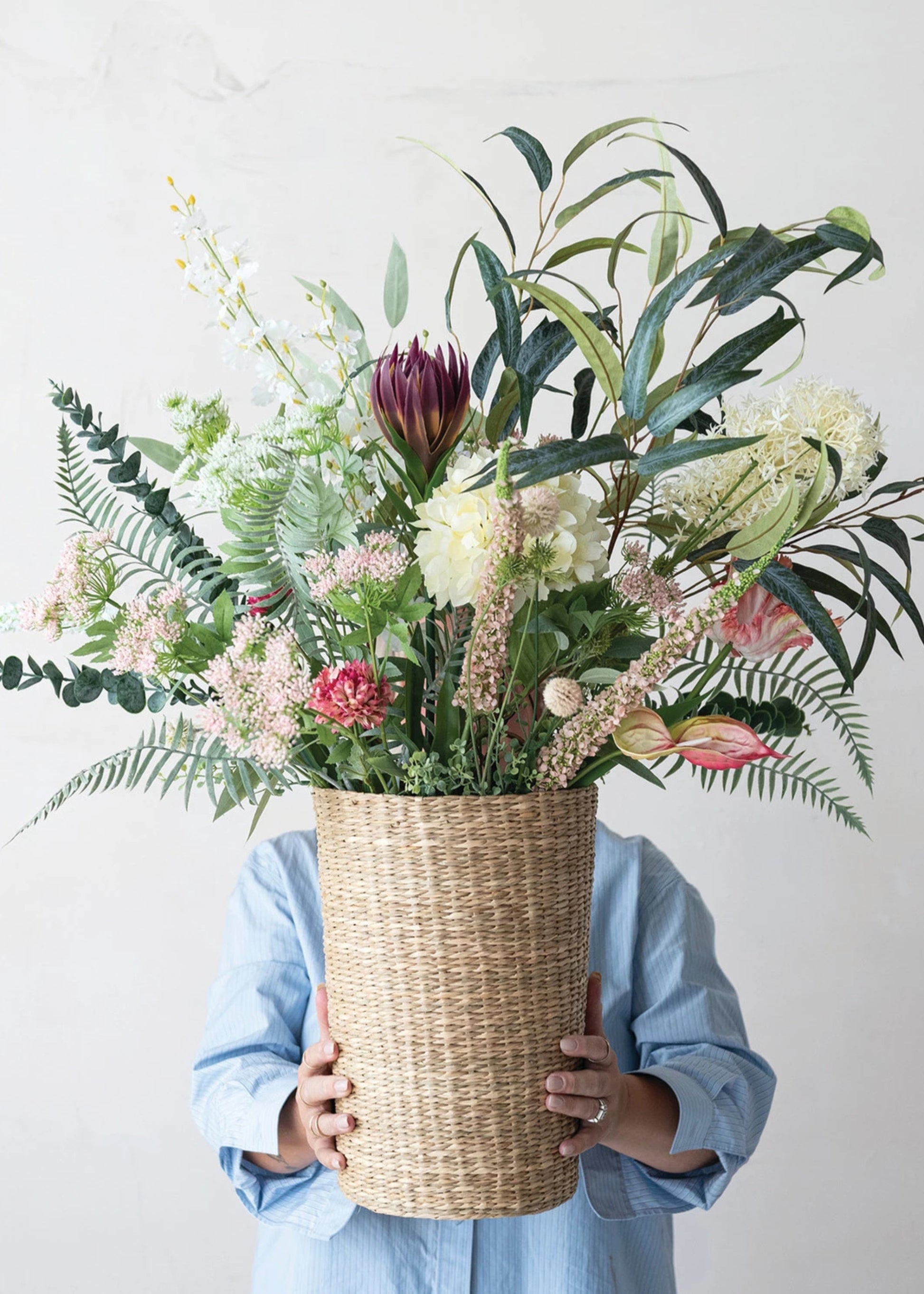 Person holding a large woven basket filled with a colorful arrangement of flowers and plants