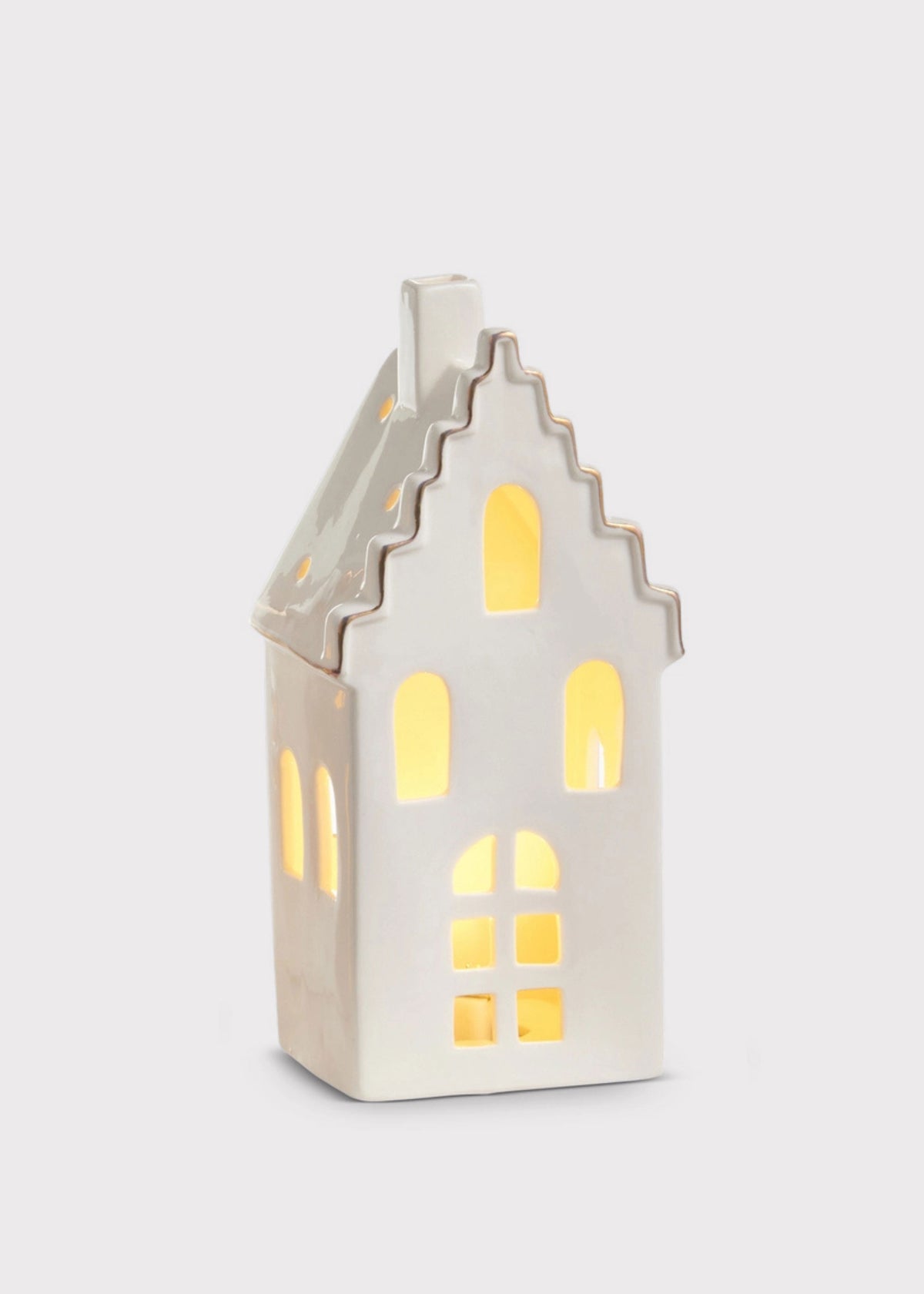 A small white ceramic house-shaped candle holder with glowing yellow window lights, set against a plain light gray background.