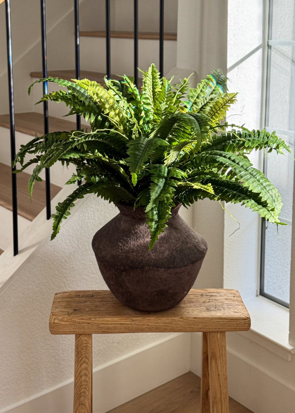 A green bostonfern plant in a dark brown textured ceramic pot, displayed on a rustic wooden stool in a bright indoor setting near a window and staircase.