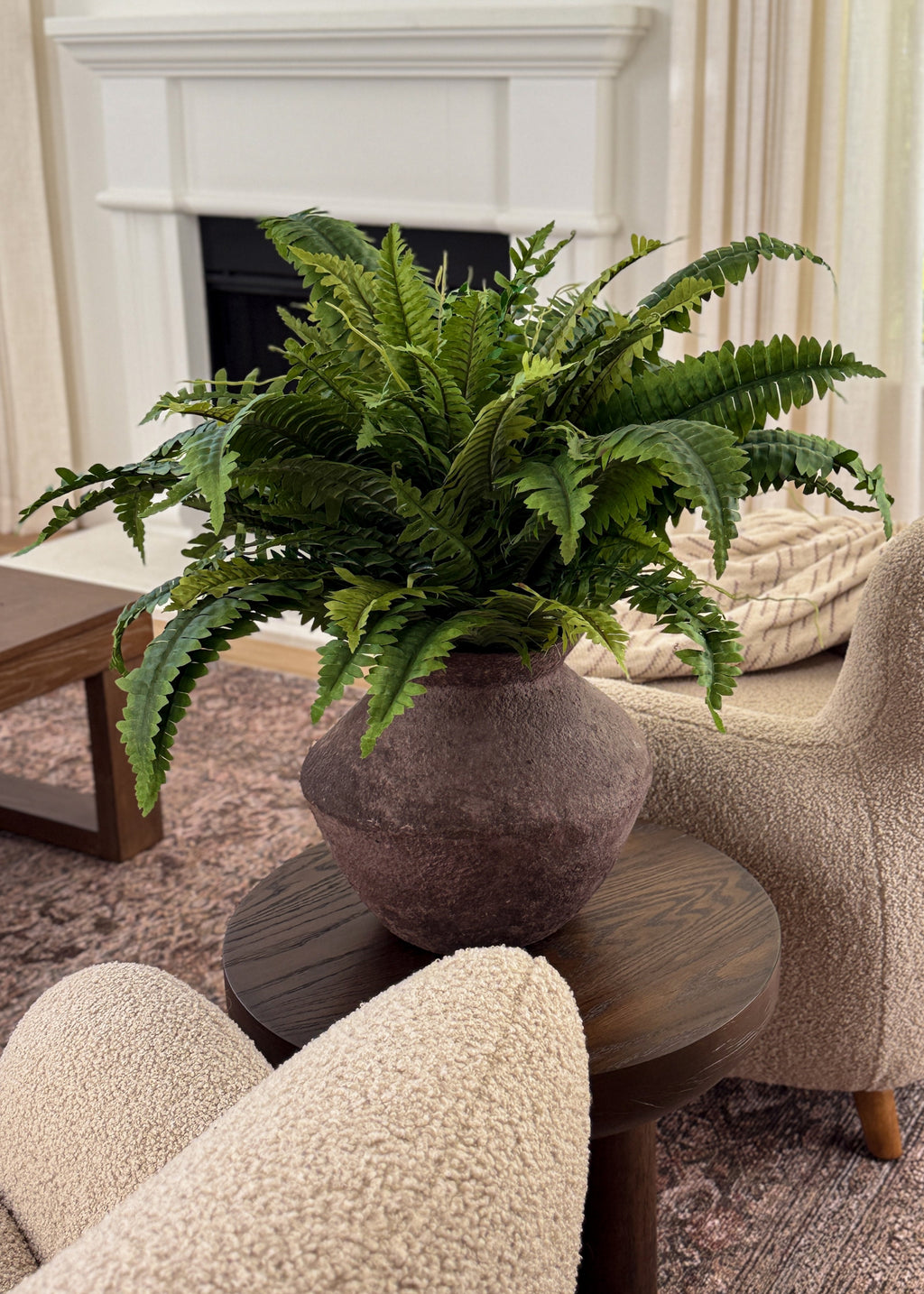 A lush green artificial boston fern in a textured brown ceramic pot, styled on a dark wood side table in a cozy living room setting with beige armchairs and a fireplace in the background.