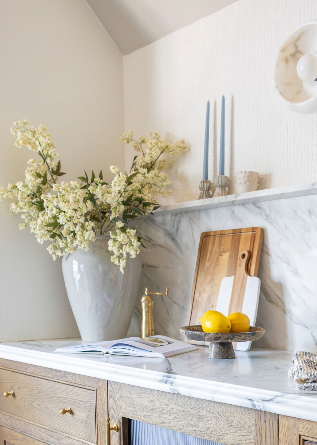 A styled interior corner with a marble countertop, light wood cabinet, white vase filled with white flowers, candles, a wooden cutting board, lemons, and an open book.