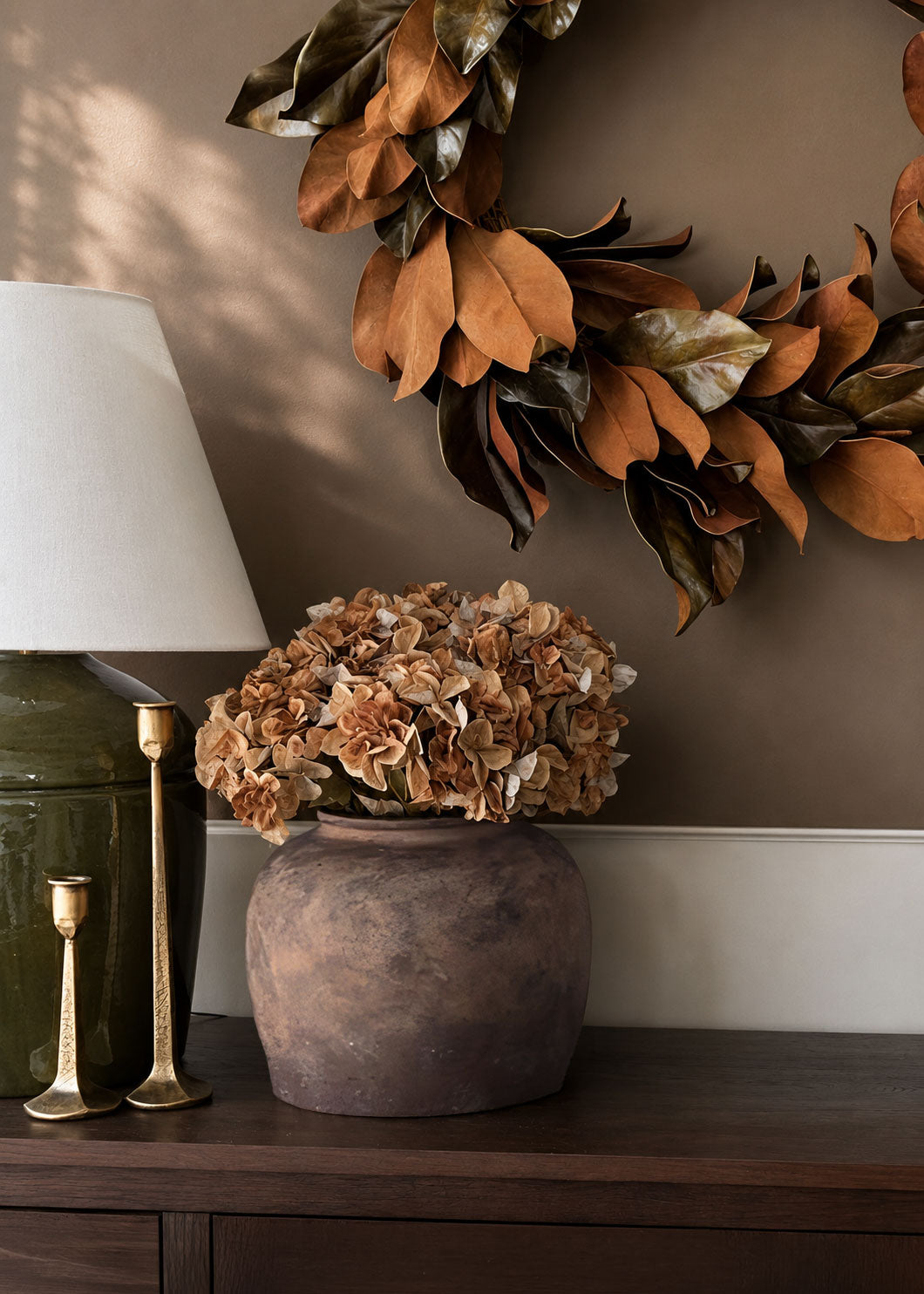 dried look artificial brown hydrangeas in a stoneware vase with a magnolia leaf wreath hanging on the wall behind