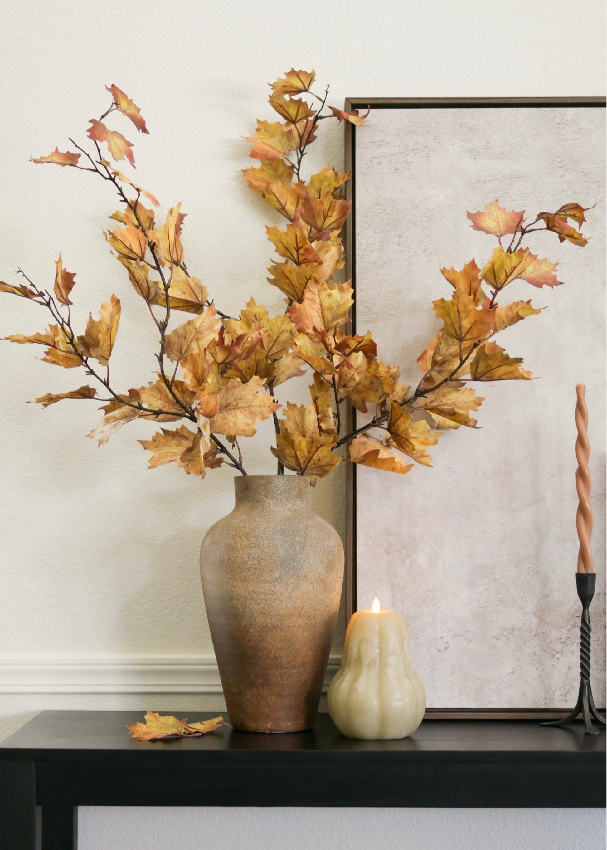 Autumn leaf artificial maple branches arranged in a rustic brown ceramic vase on a dark console table, with a small cream candle holder and a framed print in the background.
