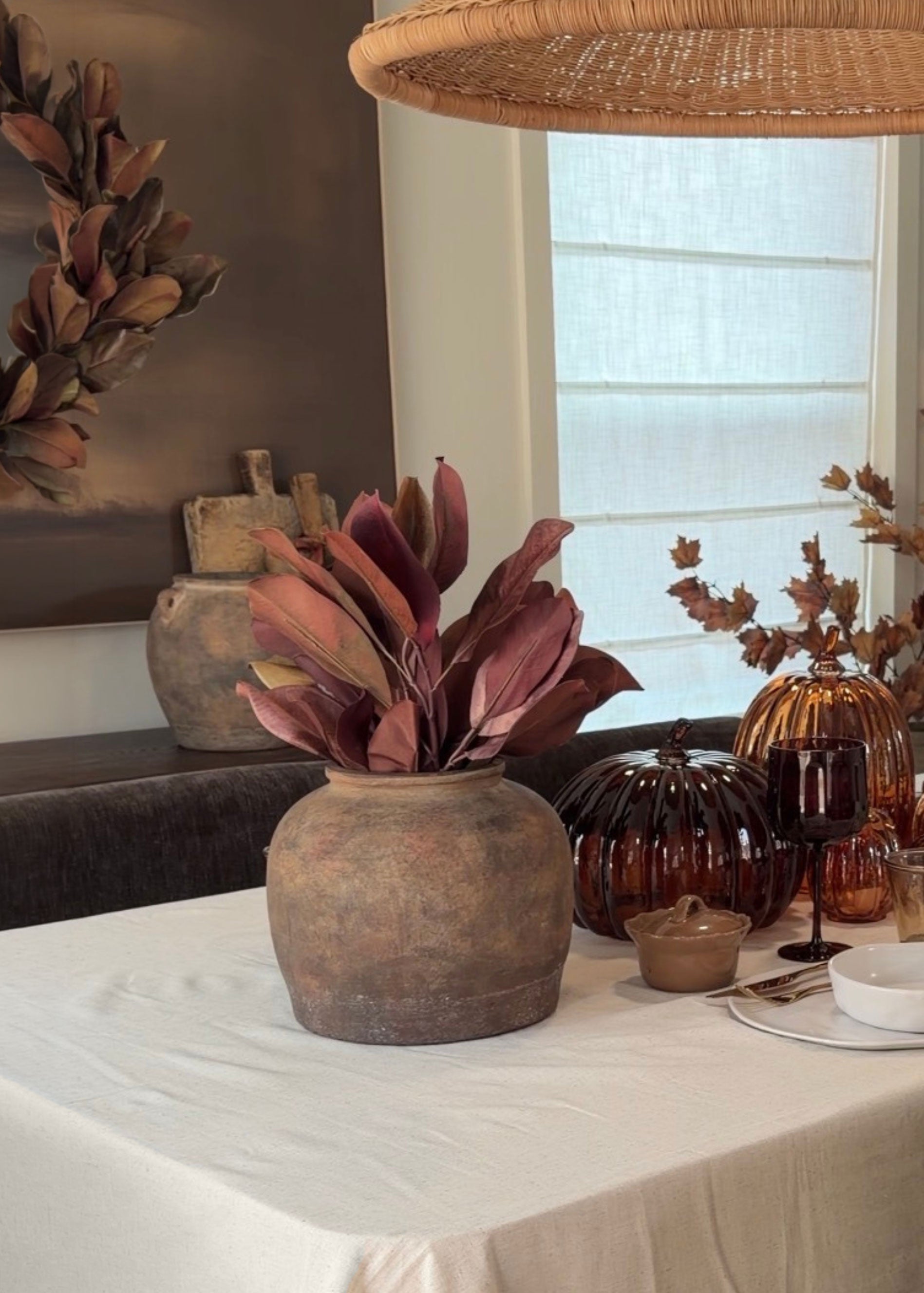 A rustic brown ceramic vase filled with burgundy brown faux magnolia leaves, styled on a white tablecloth alongside amber glass pumpkins and a white plate in a warm, decorated dining room setting.