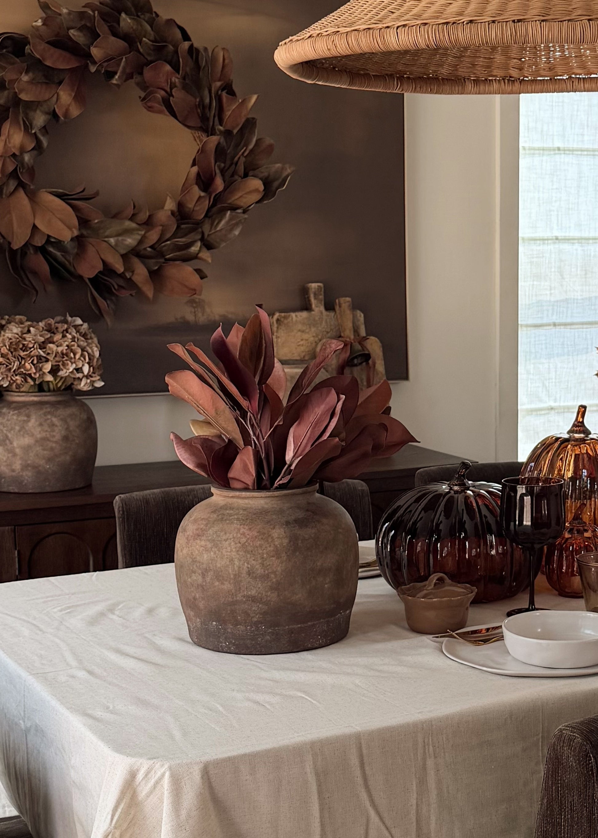 A rustic terracotta vase filled with burgundy dried artificial magnolia leaves on a white tablecloth, styled in a warm autumn dining room setting with pumpkins, a wicker lampshade, and a large magnolia leaf wreath on the wall.