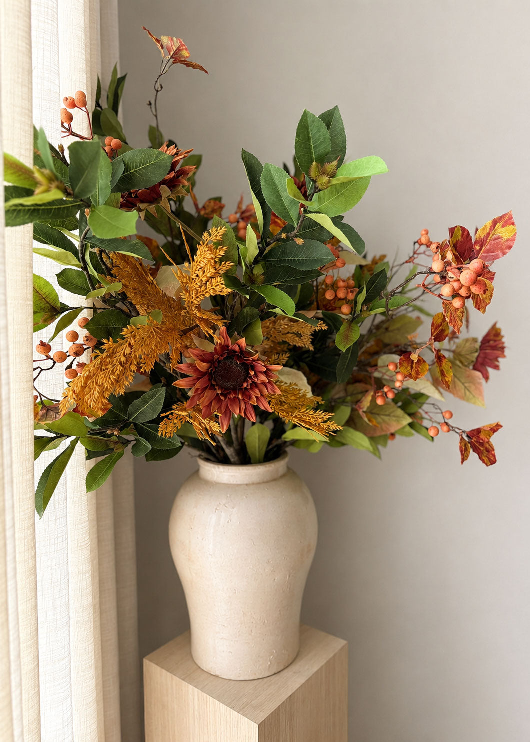 An autumn floral arrangement with green leaves, orange and red berries, and mustard-yellow textured stems, displayed in a cream ceramic vase on a light wood pedestal against a neutral wall.