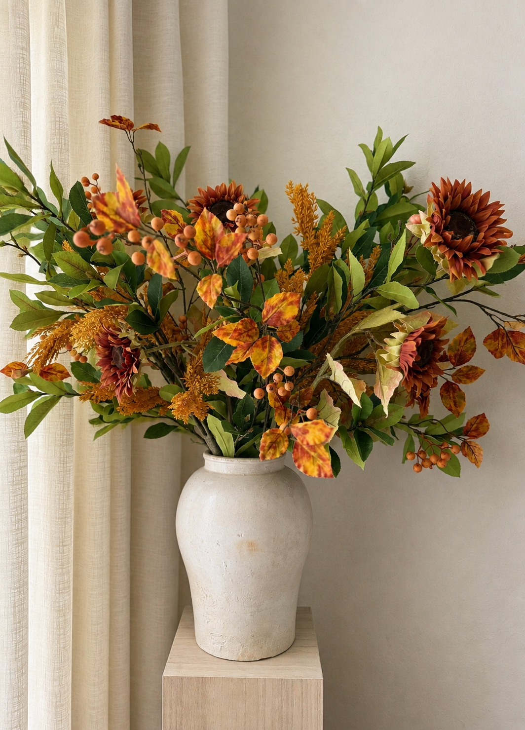 An autumn floral arrangement with sunflowers, fall leaves, and green foliage, displayed in a textured off-white ceramic vase on a light wood pedestal against a neutral background.