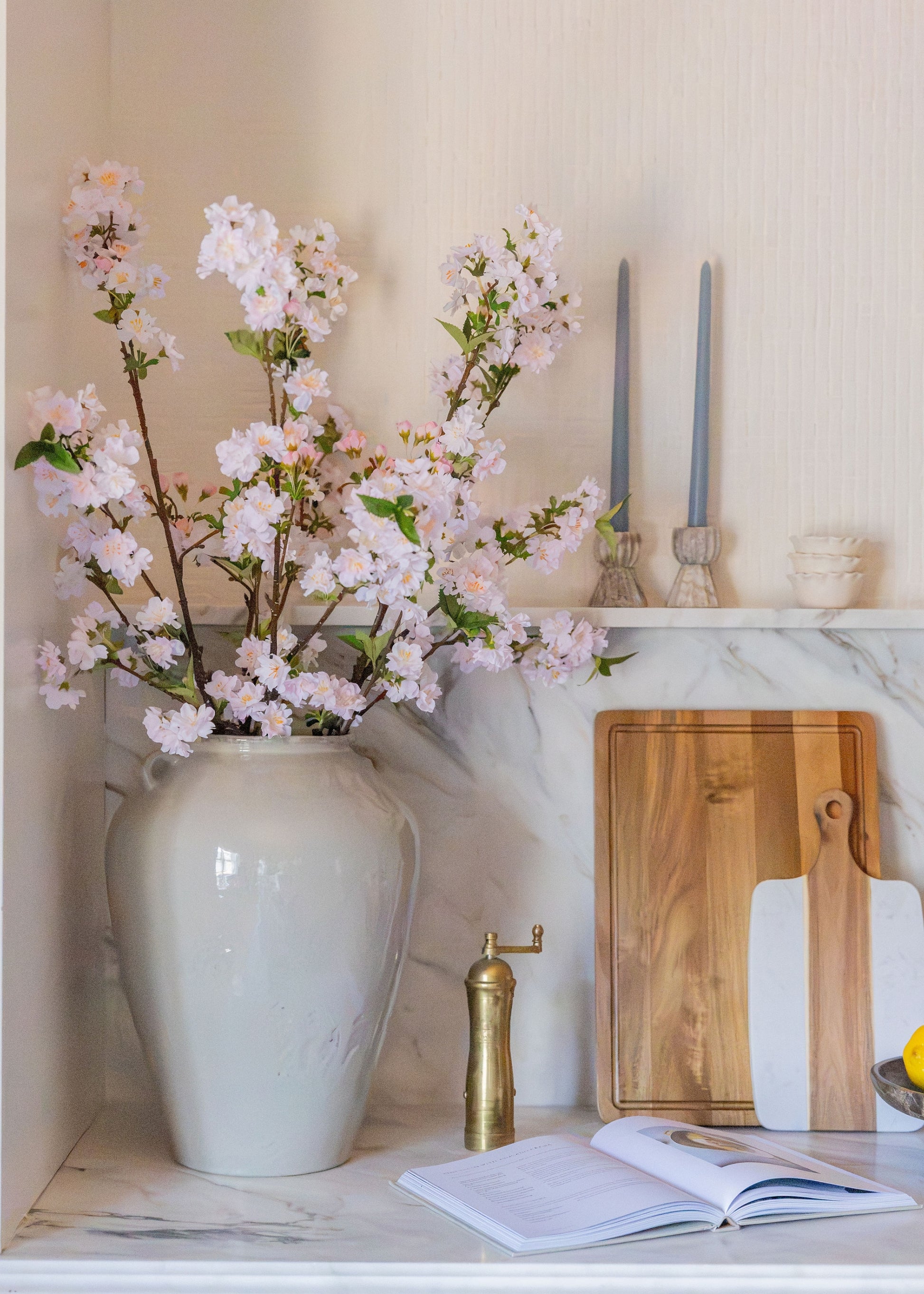 A large white ceramic vase filled with light pink cherry blossom branches, styled on a marble countertop next to wooden cutting boards, a brass pepper mill, and an open book.