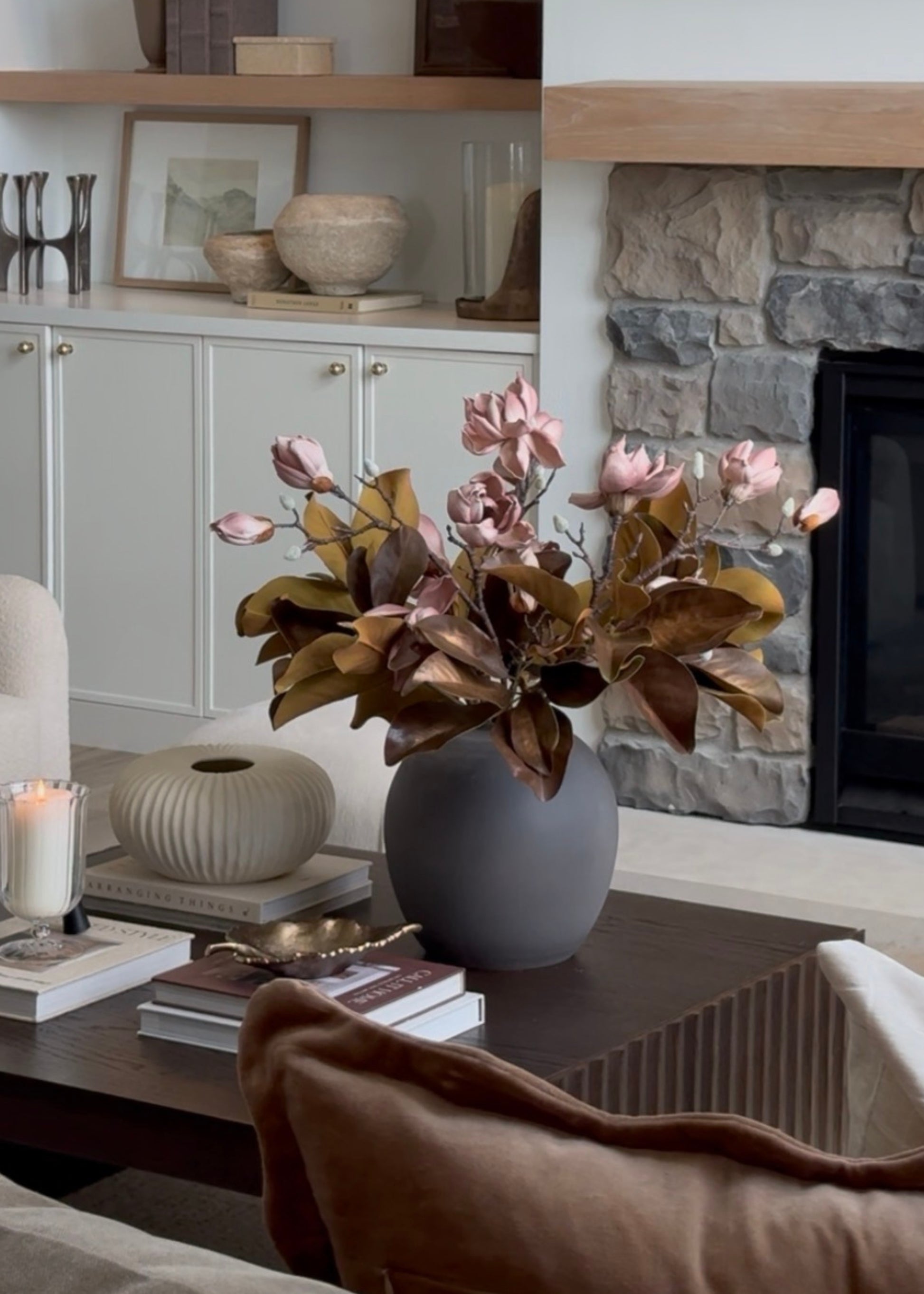 A gray ceramic vase filled with dried pink and brown magnolias on a dark wooden table in a cozy living room setting with a stone fireplace and white cabinets in the background.