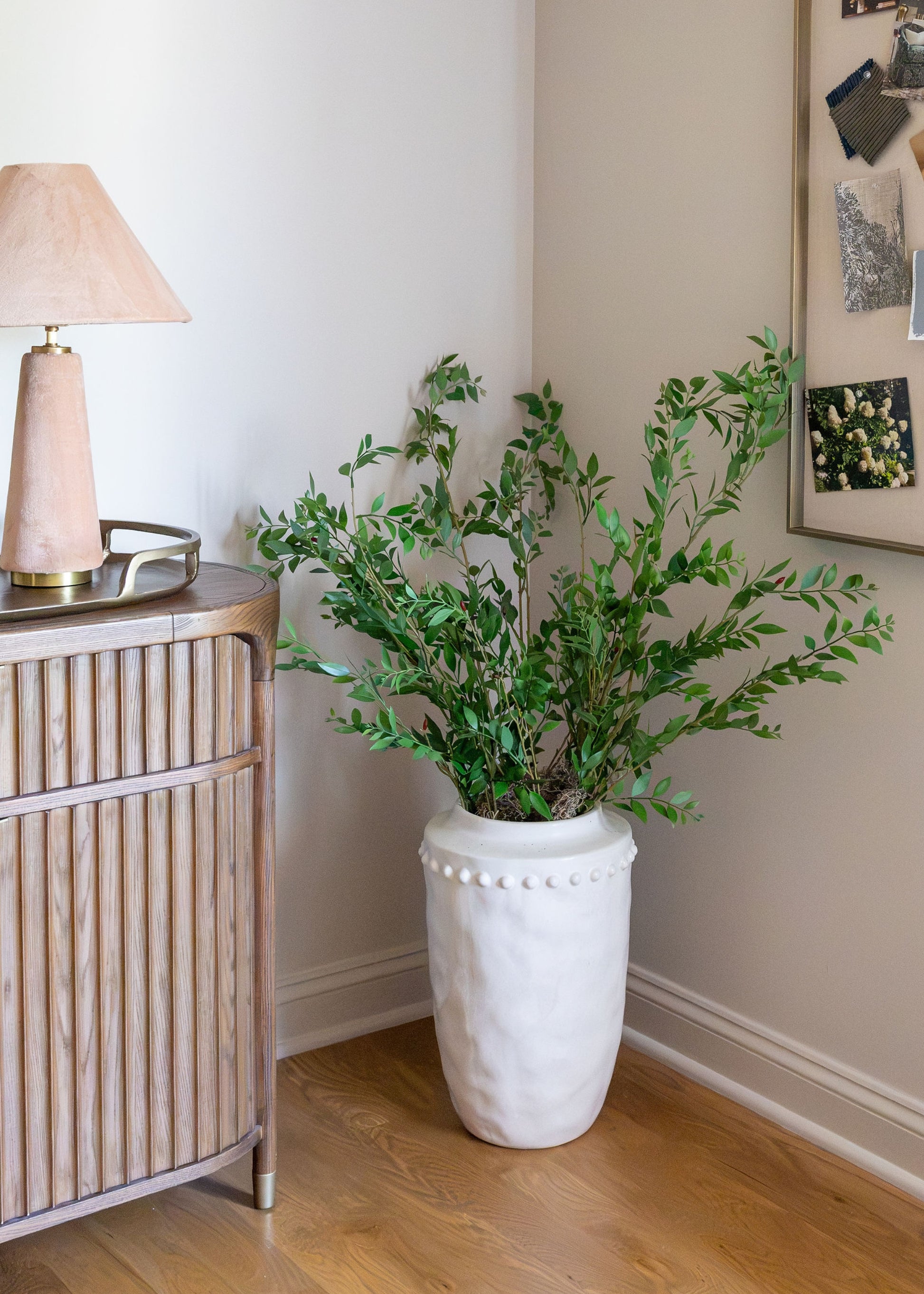 A green leafy plant in a textured white ceramic pot, styled in a corner of a room next to a light wood slatted sideboard with a lamp.