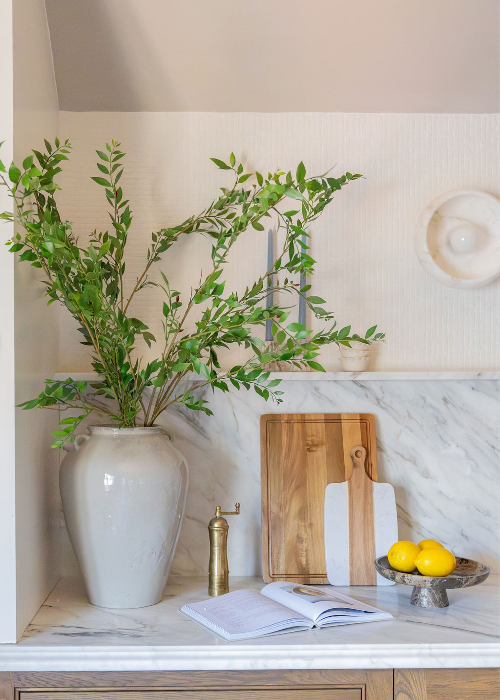 A styled kitchen countertop featuring a large white ceramic vase filled with green leafy branches, a wooden cutting board, a white-handled knife, an open cookbook, a brass pepper mill, and a bowl of lemons on a white marble surface.
