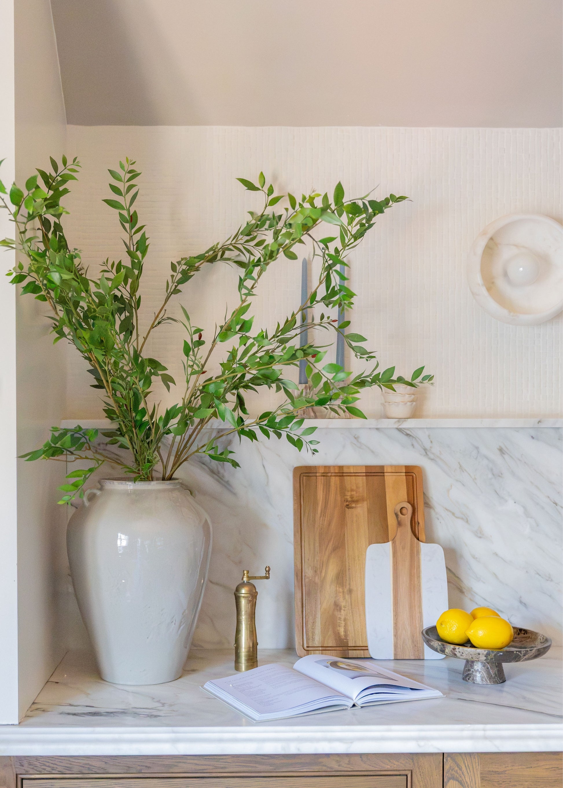 A styled kitchen countertop featuring a large white ceramic vase filled with green leafy branches, a wooden cutting board, a white-handled knife, an open cookbook, a brass pepper mill, and a bowl of lemons on a white marble surface.