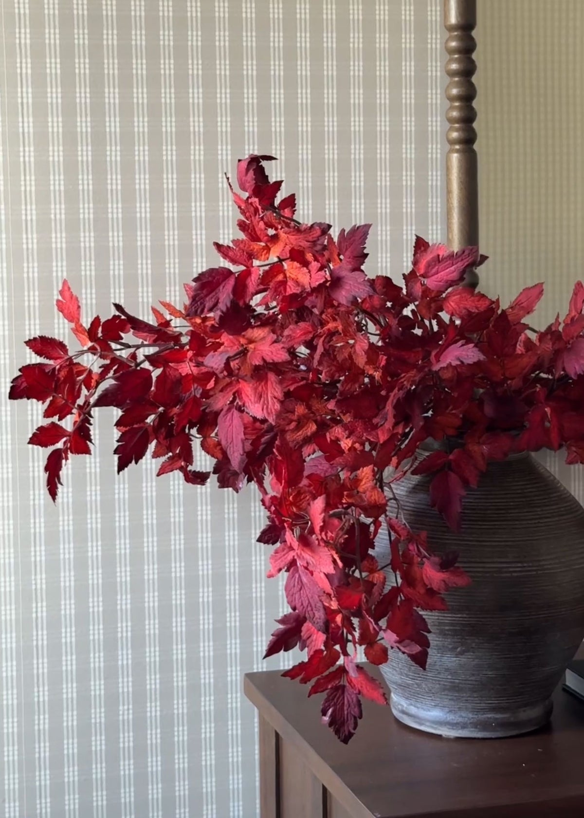 A decorative artificial autumn leaf arrangement in deep red and burgundy tones, displayed in a textured gray ceramic pot on a dark wooden side table against a striped wallpaper background.