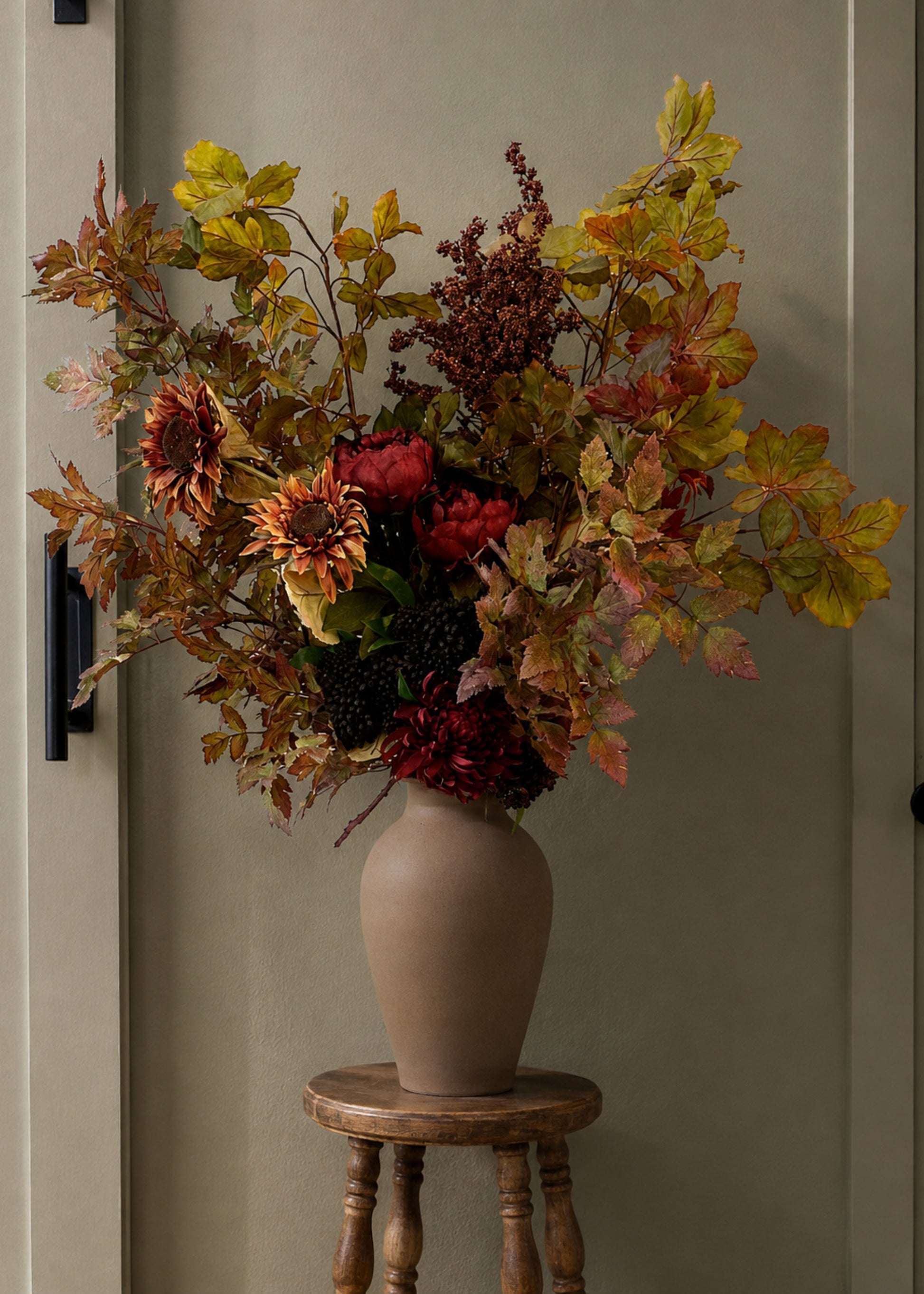 An autumn floral arrangement in a matte taupe ceramic vase, displayed on a small wooden stool against a neutral gray wall.