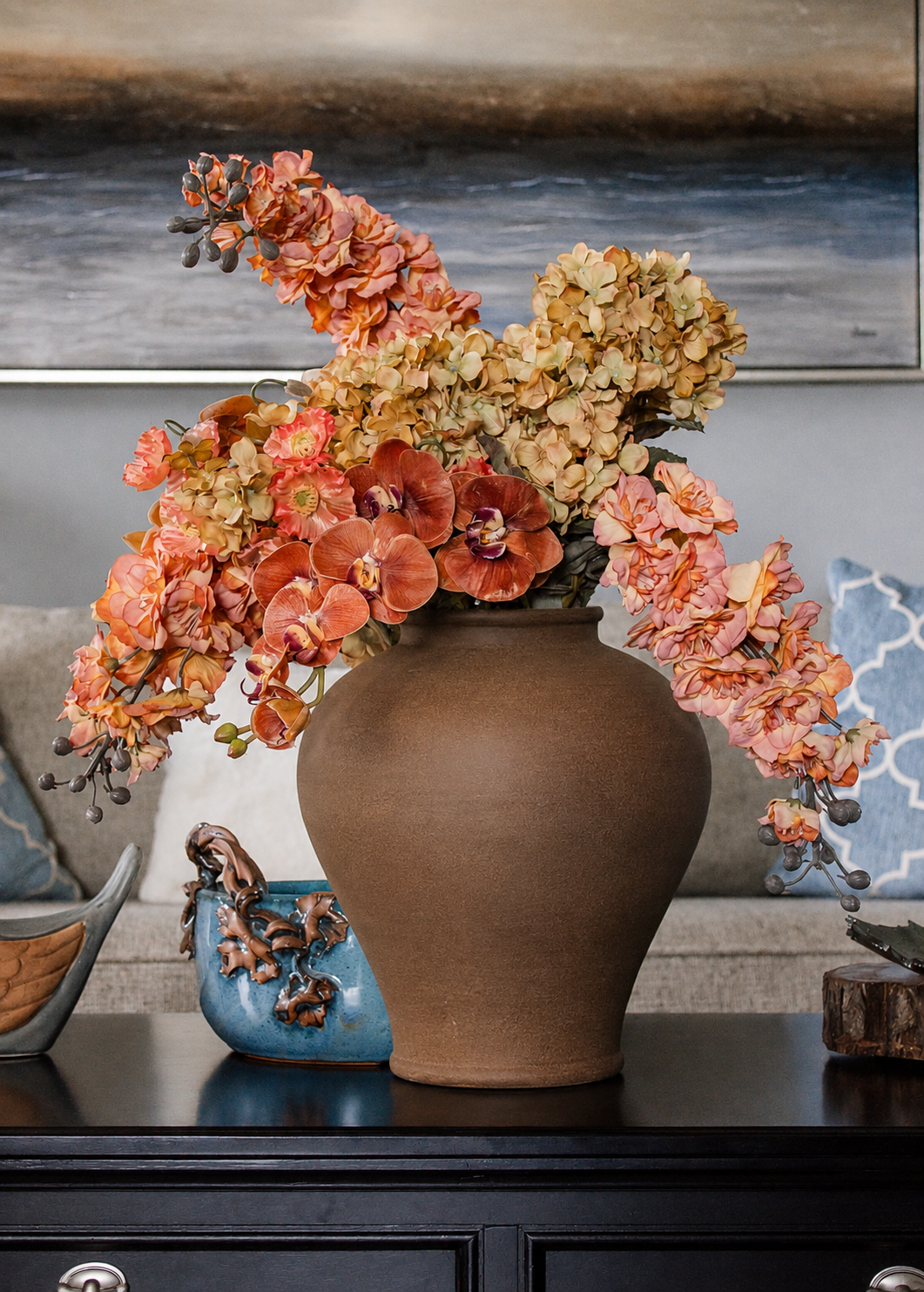 A large brown ceramic vase filled with a larkspur, hydrangea, orchid, and poppies arrangement in shades of orange, peach, and beige, displayed on a dark wooden console table in a living room setting.