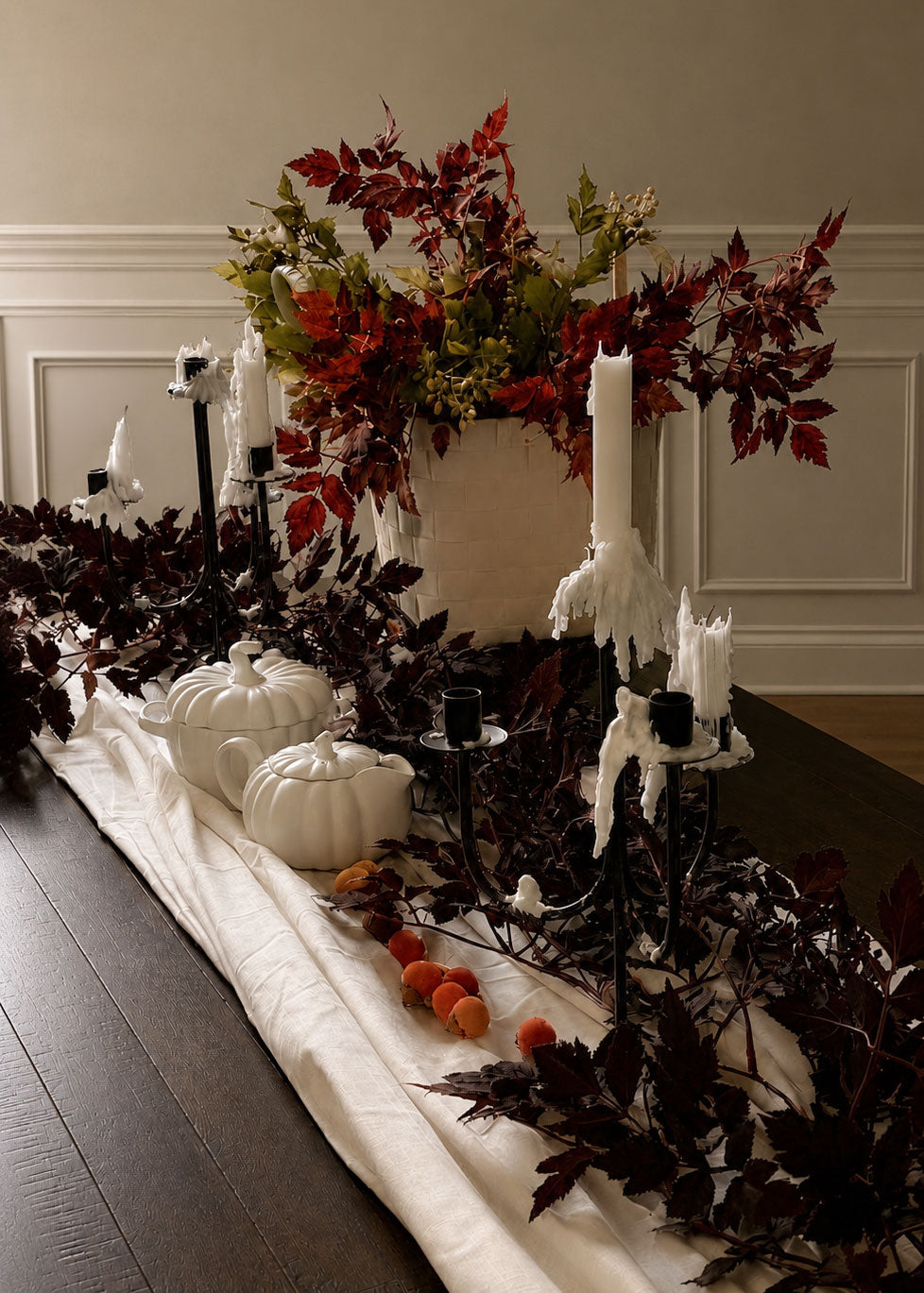 A styled autumn table centerpiece with white pumpkins, white taper candles on black candelabras, a large white basket filled with cimicifuga and Seeded Ash Leaf foliage, and a table runner decorated with a cimicifuga garland