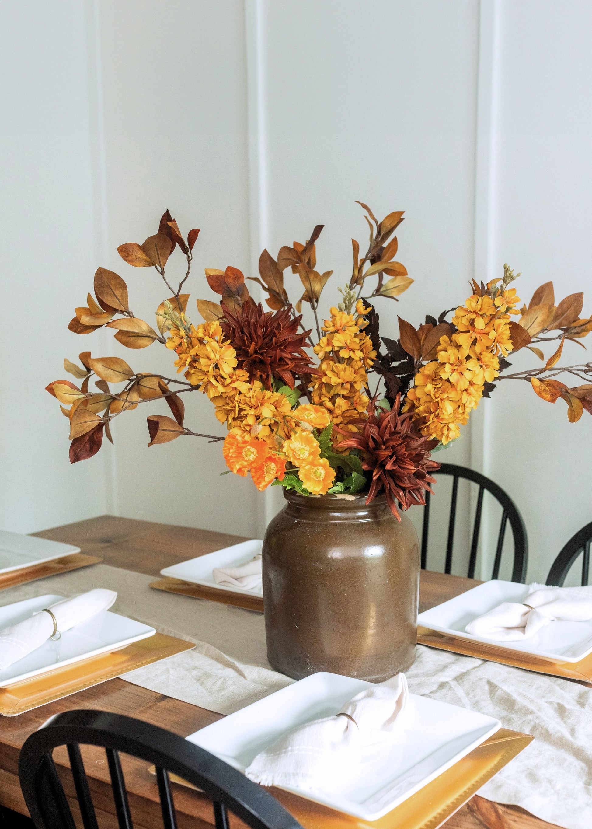 Autumn floral centerpiece in a rustic brown ceramic vase, featuring yellow and orange faux flowers with brown leaves, displayed on a wooden dining table with white plates and black chairs.